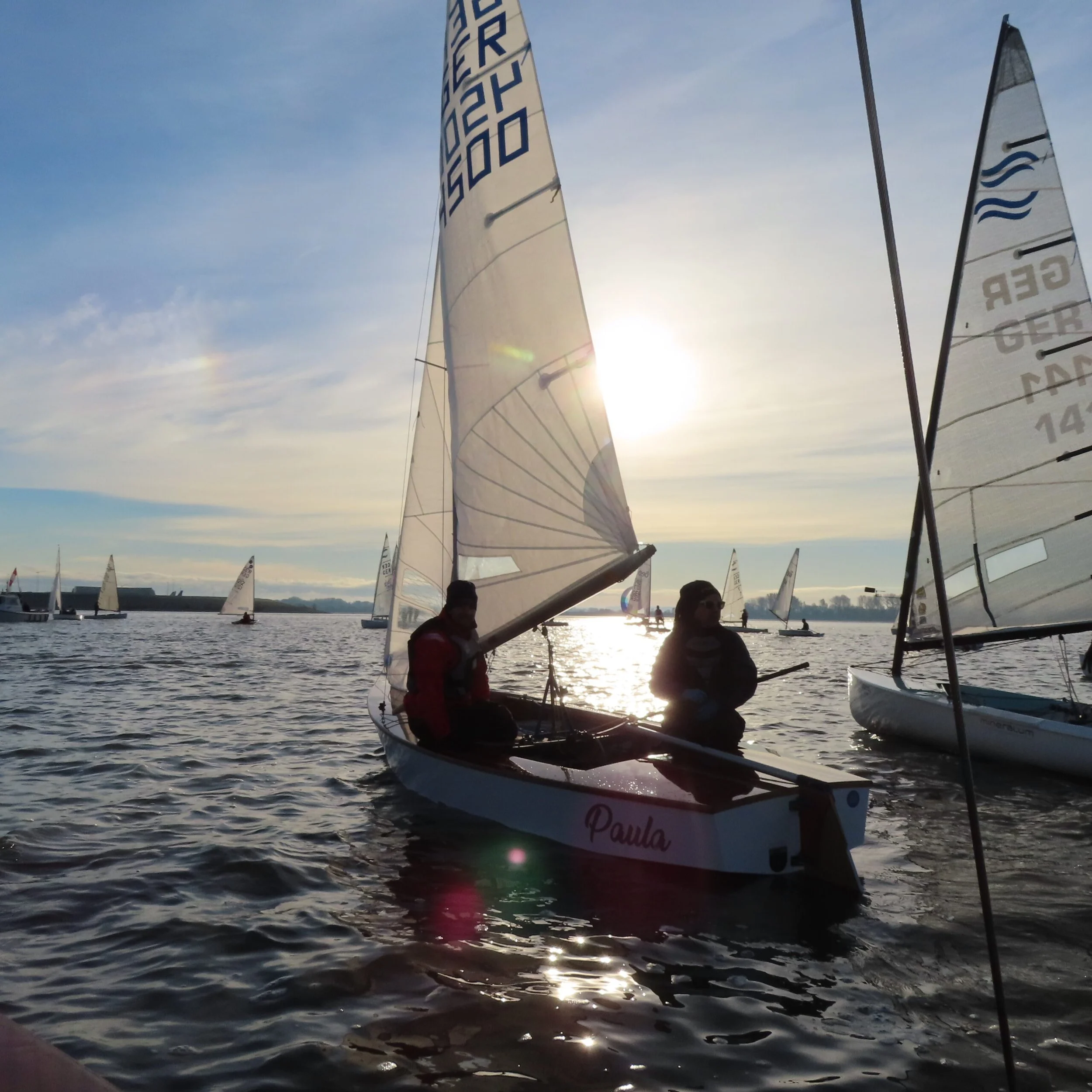 Zwei Menschen sitzen in einem kleinen Segelboot namens ‚Paula‘ auf einem Wasser bei Sonnenuntergang, umgeben von mehreren Segelbooten, darunter auch andere Boote mit weißen Segeln. Der Himmel ist teilweise bewölkt, und die Sonne ist fast untergegange