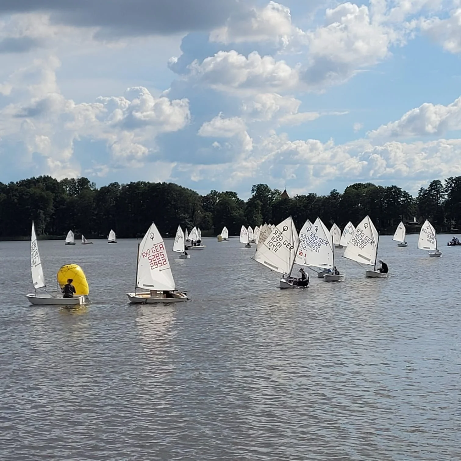 Segelboote auf einem See mit weißen Segeln und einem gelben Ball auf einem Boot, bei bewölktem Himmel.