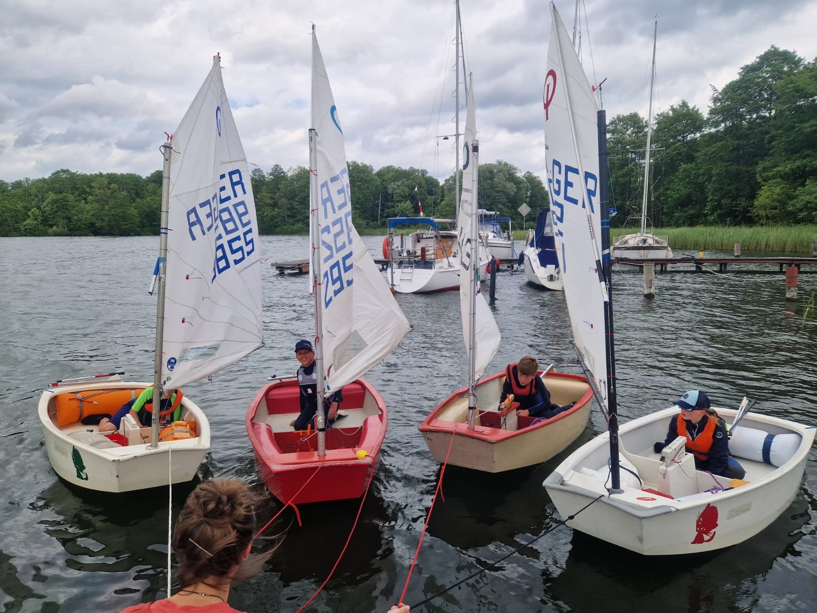 Vier kleine Segelboote mit Kindern, die auf einem ruhigen See segeln, während eine Erwachsene im Vordergrund das Geschehen beobachtet.