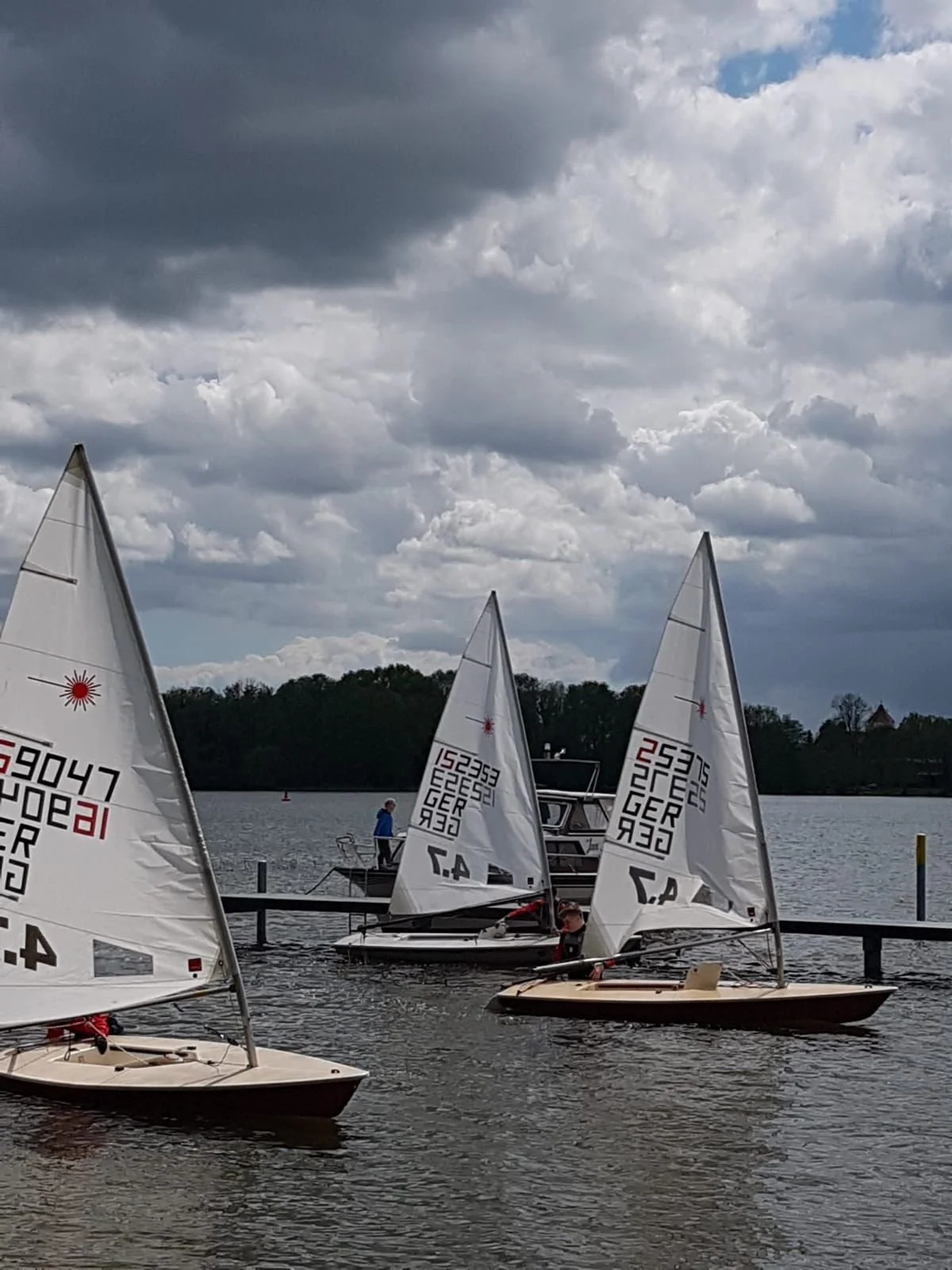 Drei kleine Segelboote liegen im Wasser an einem Steg auf einem See, mit dunklen Wolken am Himmel, im Hintergrund Bäume und ein Haus auf einem Hügel.