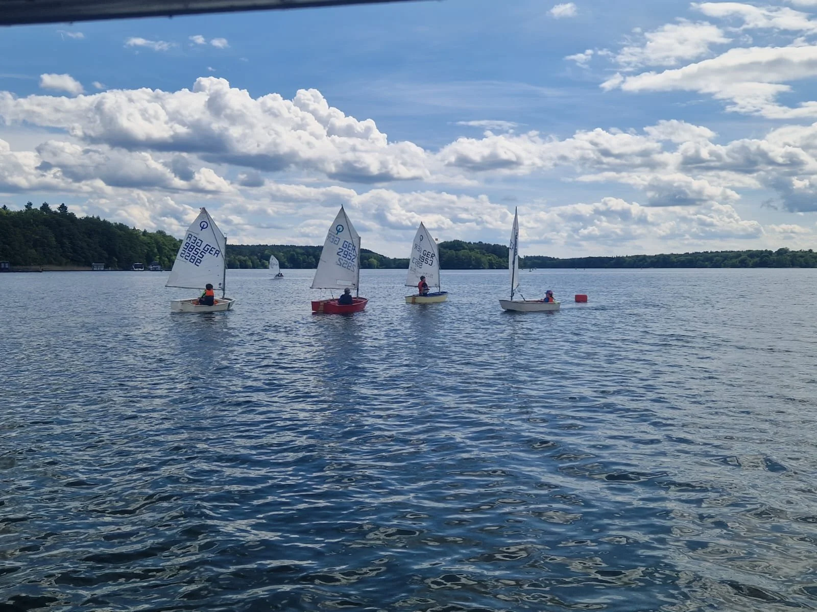 Vier kleine Segelboote auf einem ruhigen See bei sonnigem Himmel mit Wolken, umgeben von Wald.