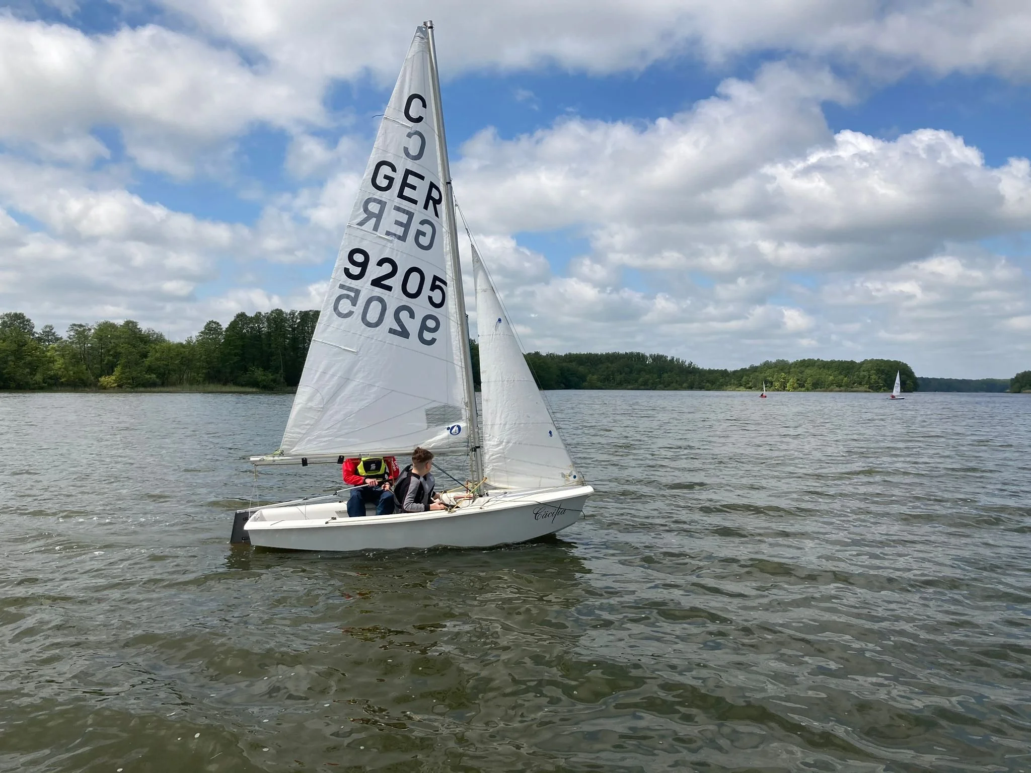 Zwei Menschen segeln auf einem kleinen weißen Boot auf einem ruhigen See mit bewölktem Himmel und Bäumen im Hintergrund.