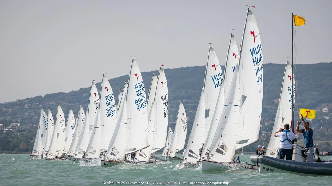 Segelboote bei einem Rennen auf einem See, mit einem Begleitschiff und Windfahnen, im Hintergrund Hügel