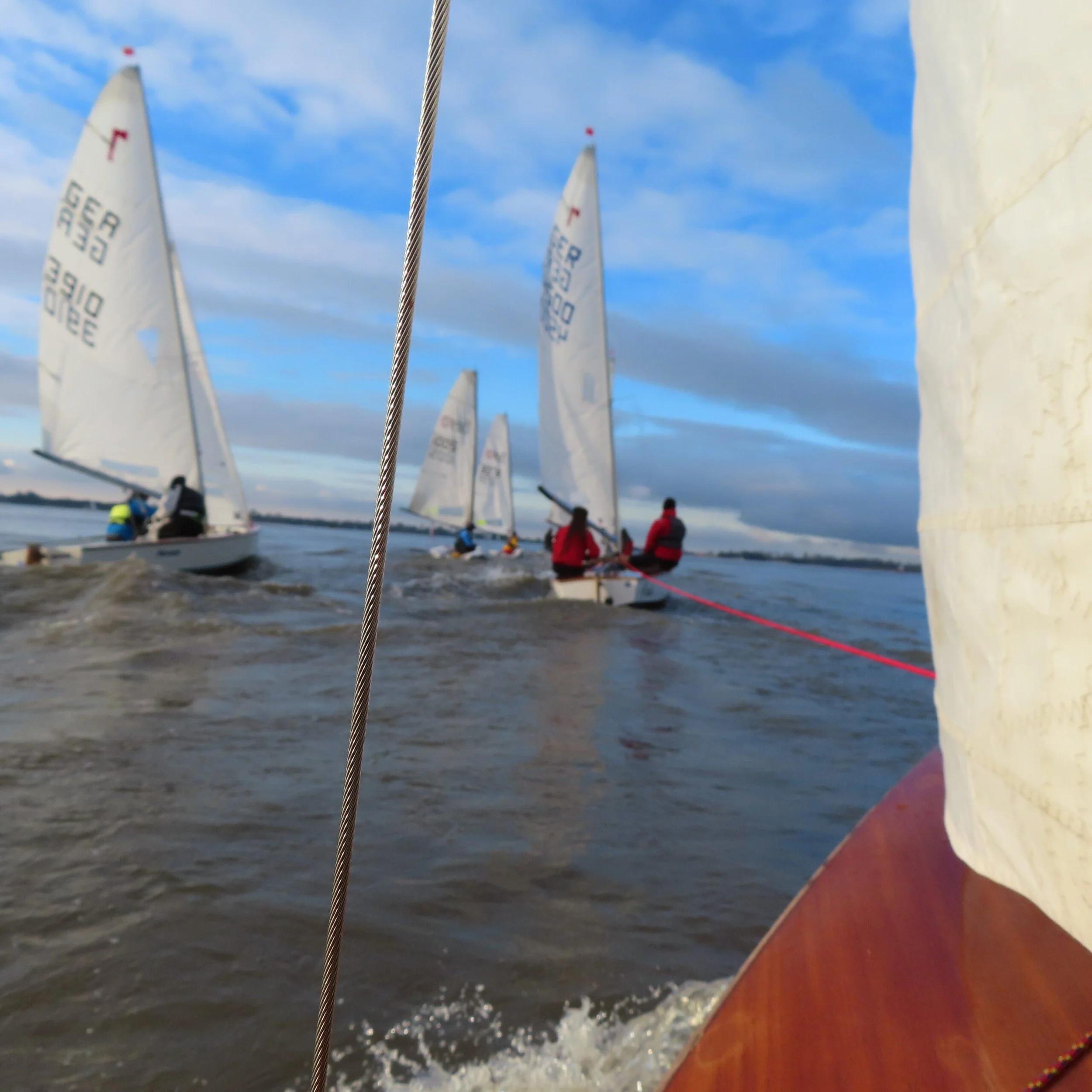 Segelboote auf dem Wasser bei Segelregatta, fotografiert vom Boot im Vordergrund, mit mehreren Booten in der Entfernung unter einem bewölkten Himmel.