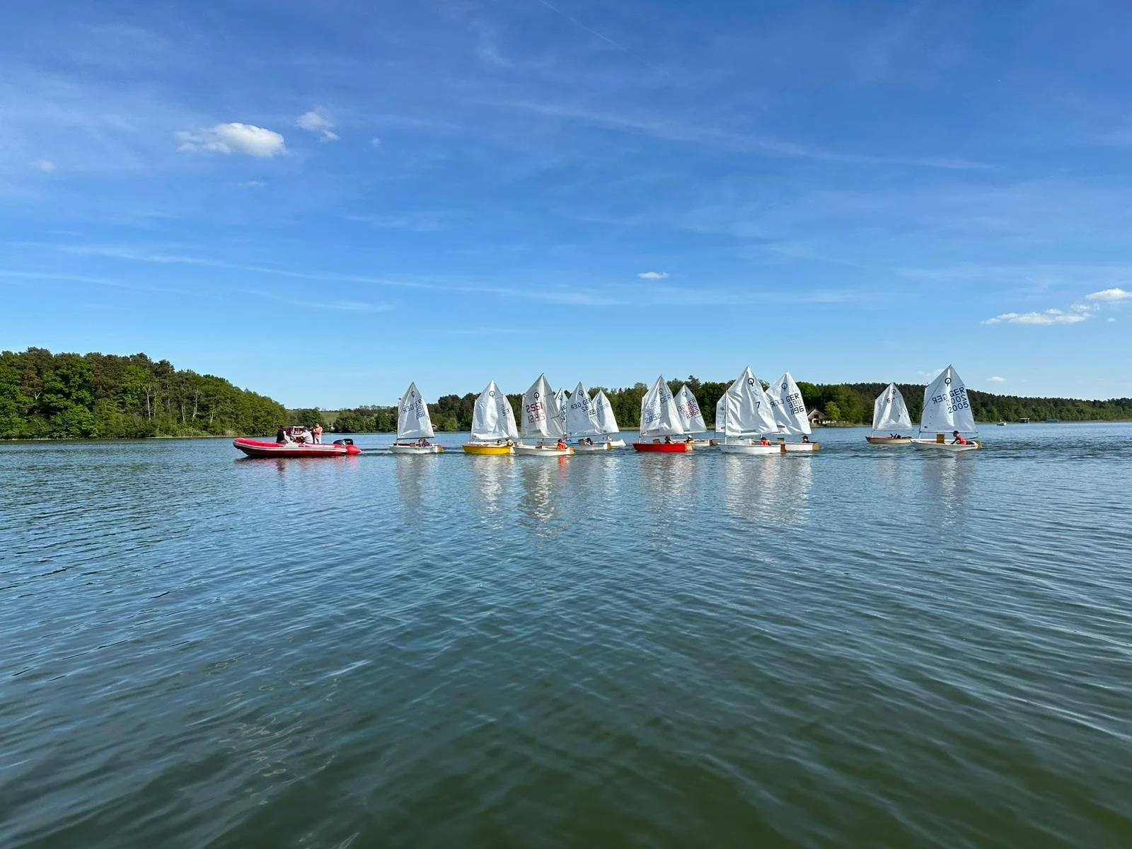 Mehrere Kinder und Jugendliche segeln mit kleinen Booten auf einem ruhigen See bei sonnigem Wetter, im Hintergrund sind Bäume und ein blauer Himmel mit wenigen Wolken.