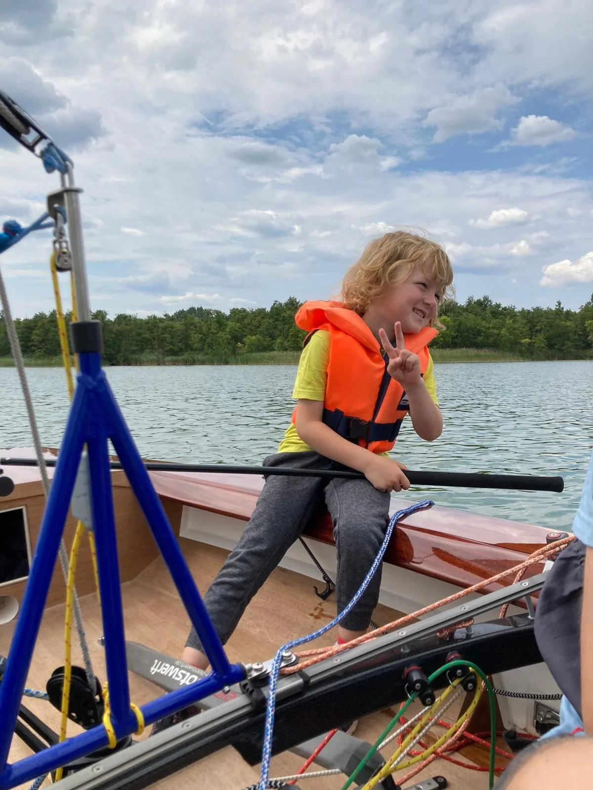 Ein Kind in einem gelben T-Shirt und einer orange Rettungsweste, das auf einem Boot auf einem See sitzt, winkt und macht einen peace Zeichen mit den Fingern, im Hintergrund Himmel und Bäume.