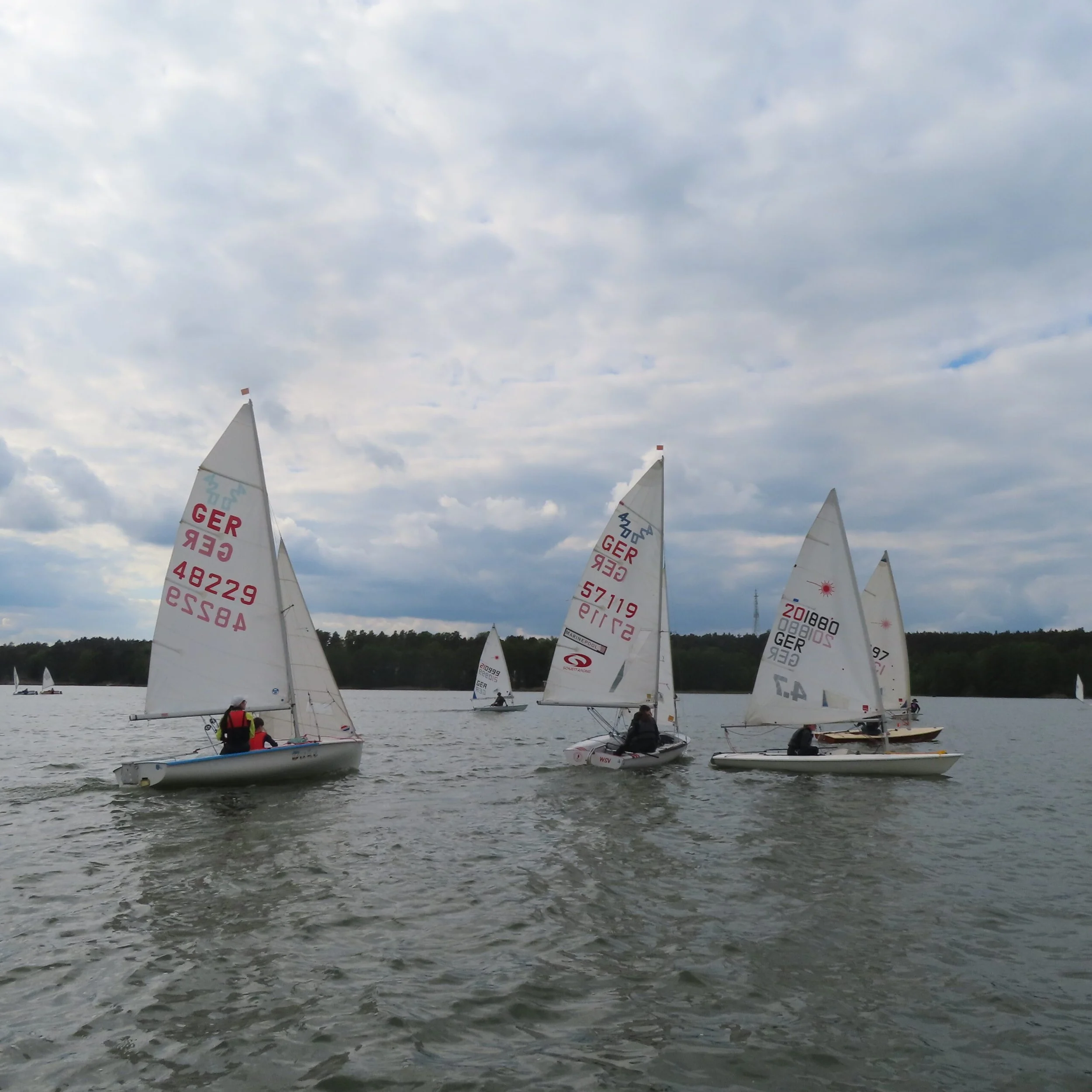 Segelboote auf einem See bei bewölktem Himmel, einige Boote in Bewegung, andere liegen ruhig, im Hintergrund Bäume am Ufer.