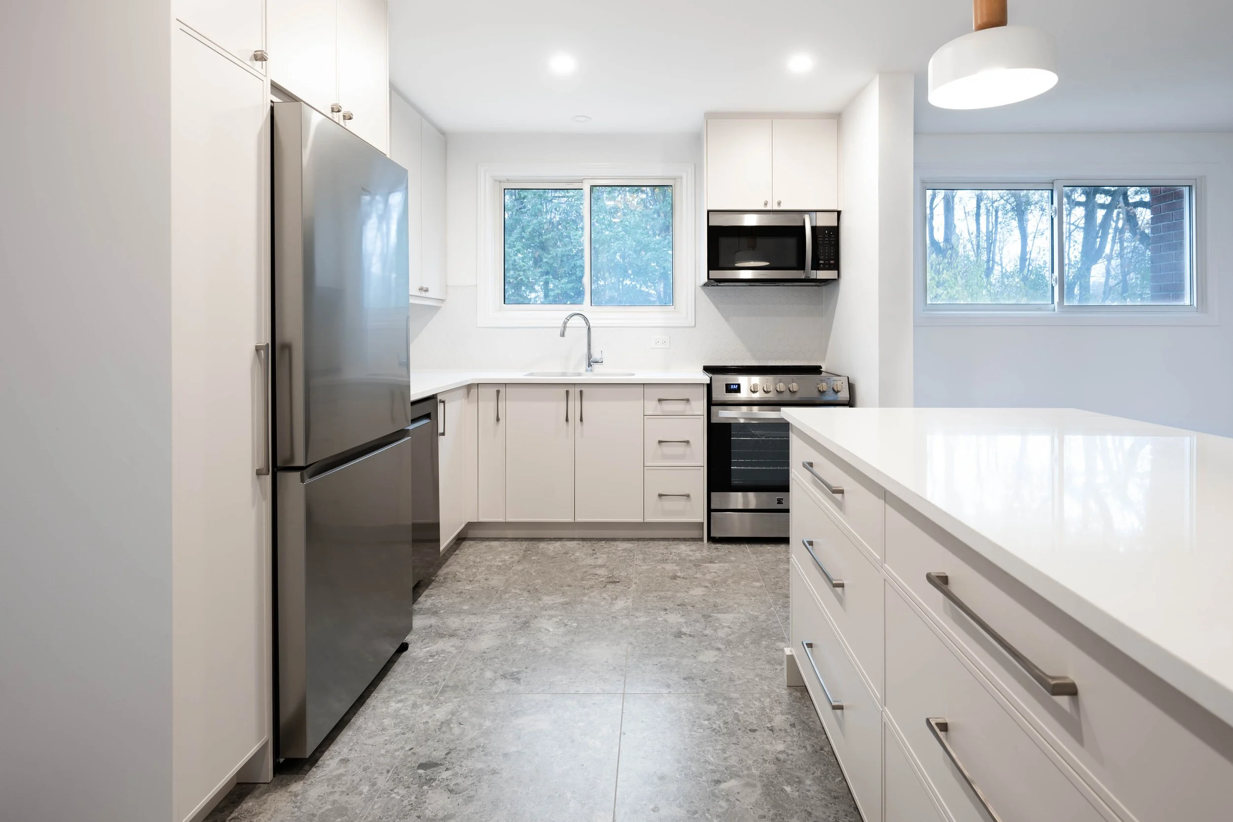 Modern kitchen with white cabinets, stainless steel refrigerator, oven, microwave, and a large kitchen island, with windows showing trees outside.
