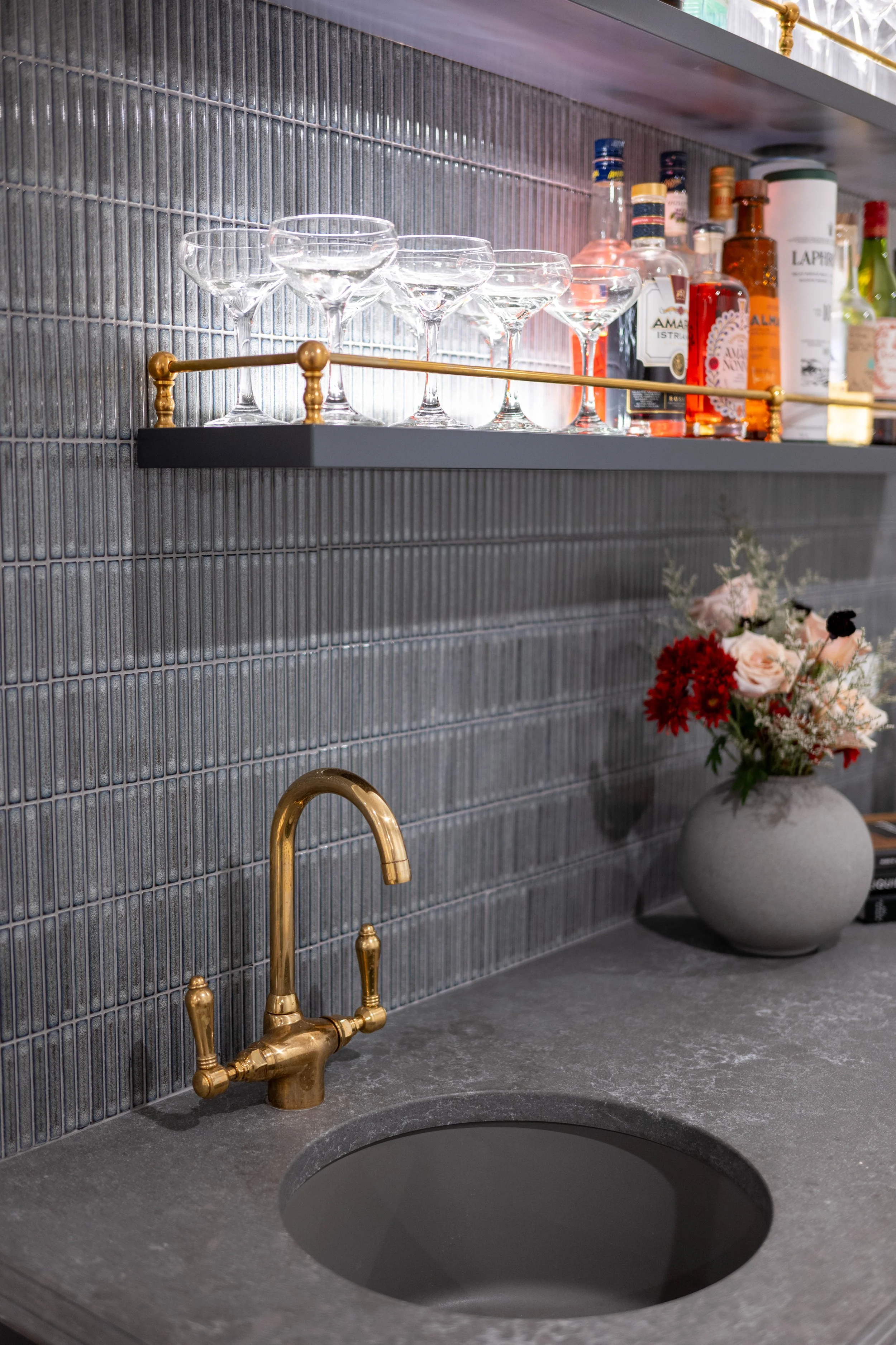 A kitchen sink with a brass faucet on a gray stone countertop, a shelf with empty glasses and bottles, and a vase with flowers against a textured gray tiled wall.