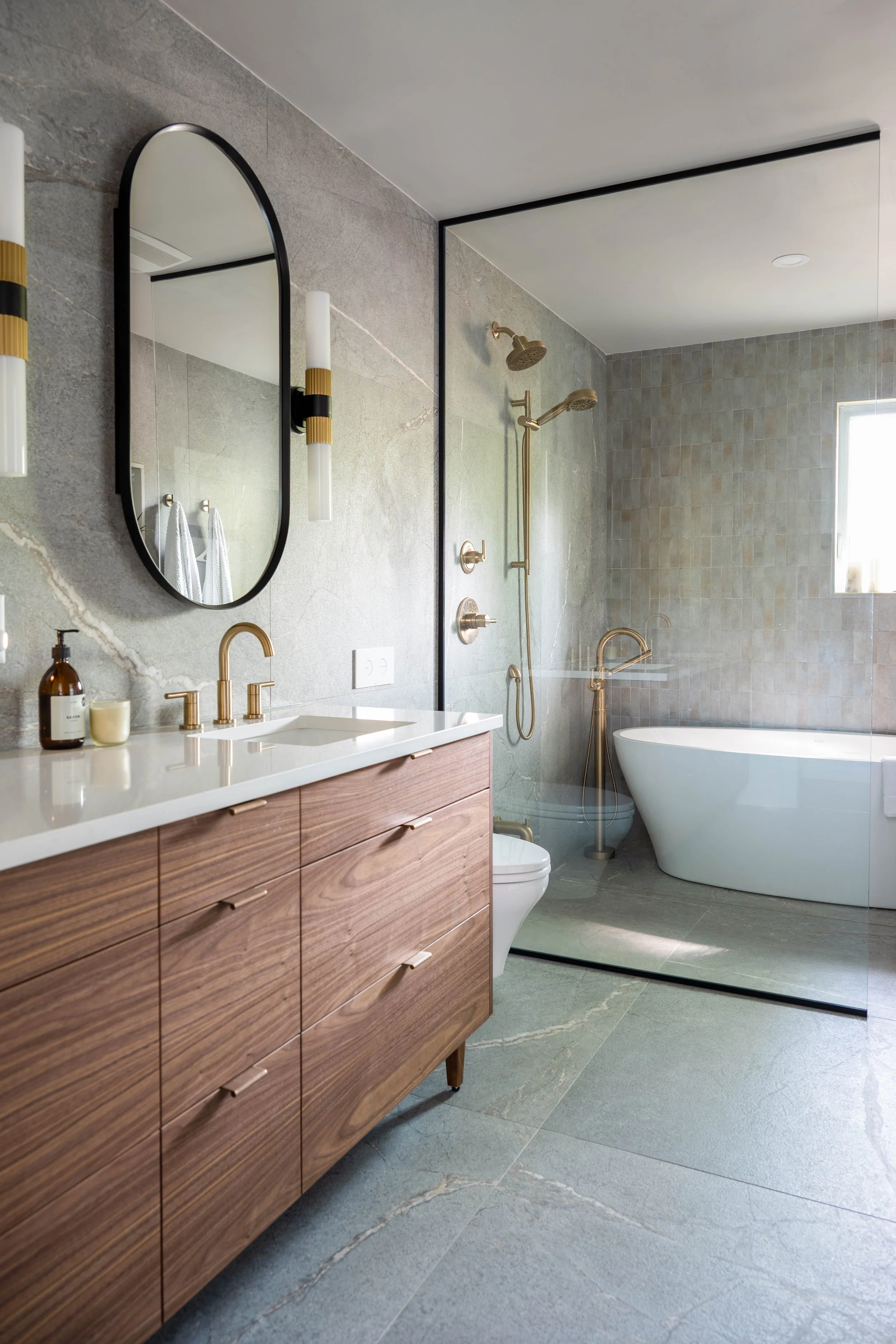 Modern bathroom with a wooden vanity, white countertop, oval mirror, brass fixtures, glass-enclosed shower, and a freestanding bathtub near a window.