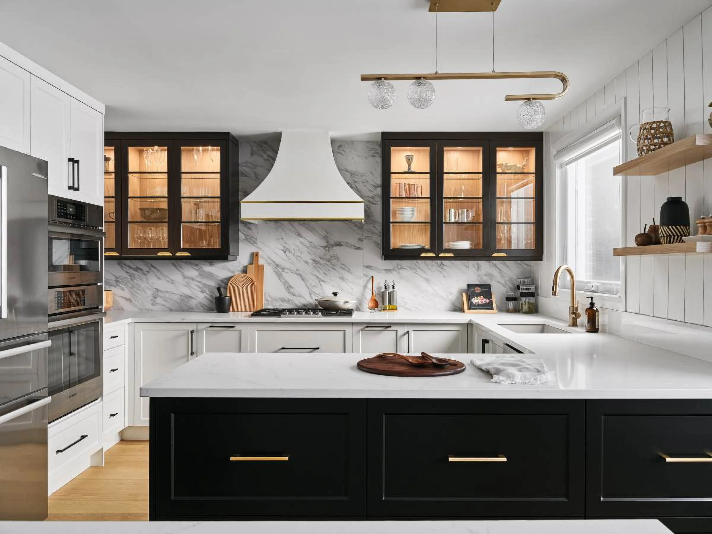 Modern kitchen with white and black cabinetry, marble backsplash, and gold accents. There is a central white island with black drawers, open wooden shelves with decor, and glass-front cabinets with dishes and glassware.