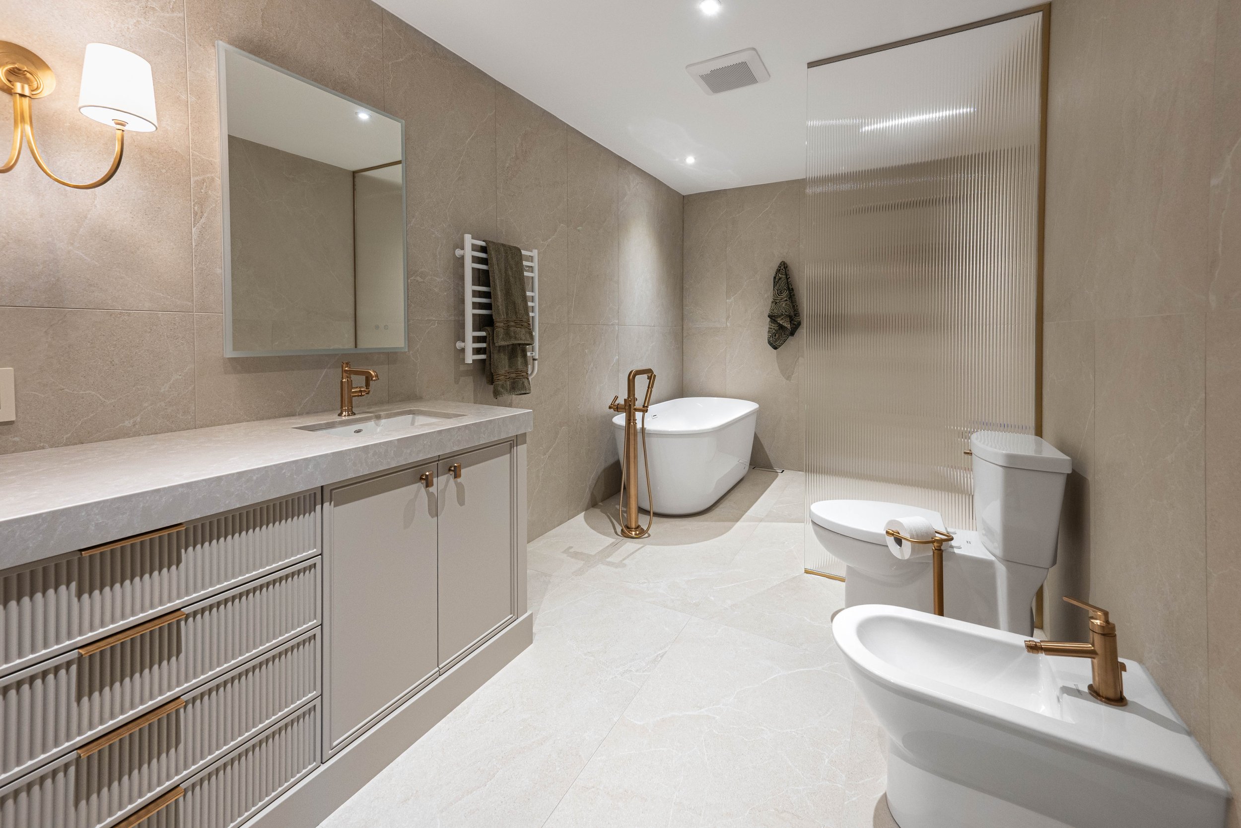 Modern bathroom with beige tiled walls and floor, featuring a vanity with a sink and mirror, a bathtub with a gold stand, a toilet, a bidet, and a textured gold-colored divider. Towel rack and towels are also visible, with warm lighting.