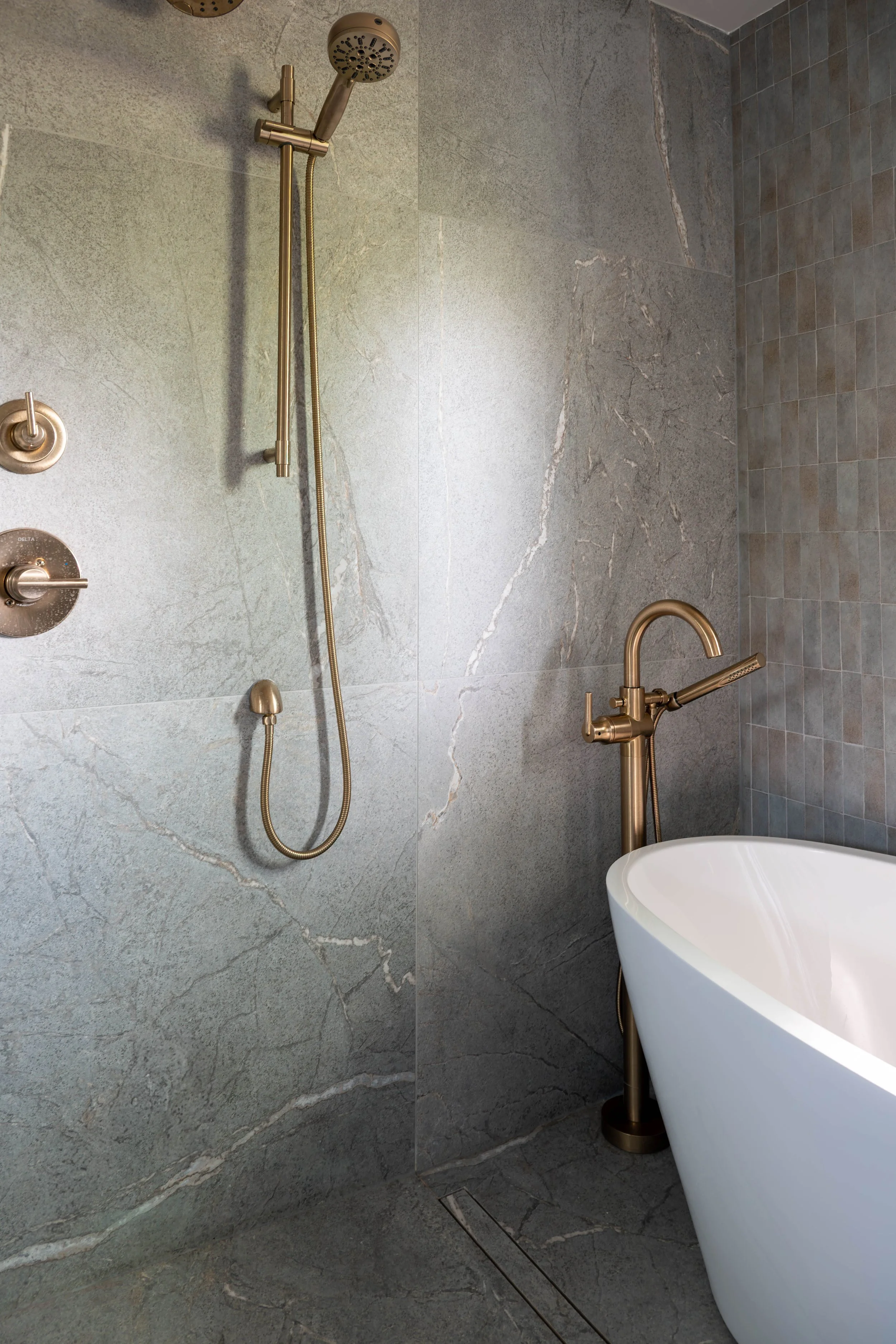 A modern bathroom with a walk-in shower featuring a gold showerhead and controls, a handheld shower, and a freestanding bathtub with a gold faucet. The walls and floor are covered in gray stone tiles.