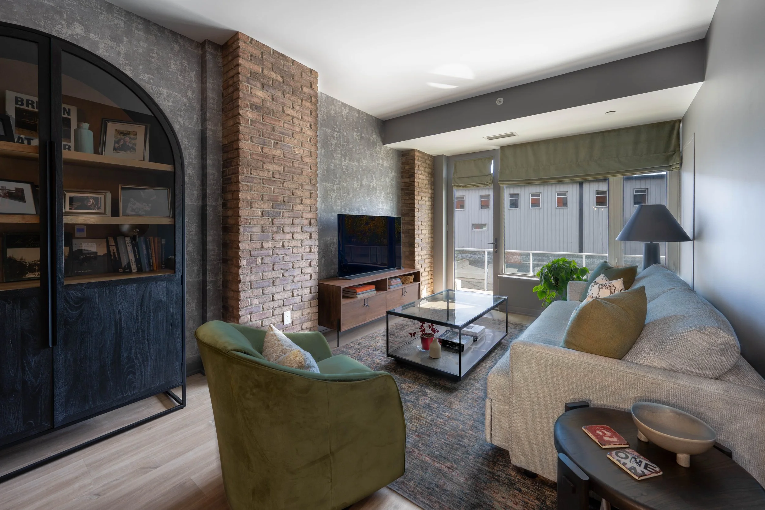 Living room with brick and concrete walls, a beige sofa with yellow pillows, a green armchair, a glass coffee table, a TV on a wooden stand, large windows with green window coverings, a bookshelf on the left side, and decorative items such as a plant