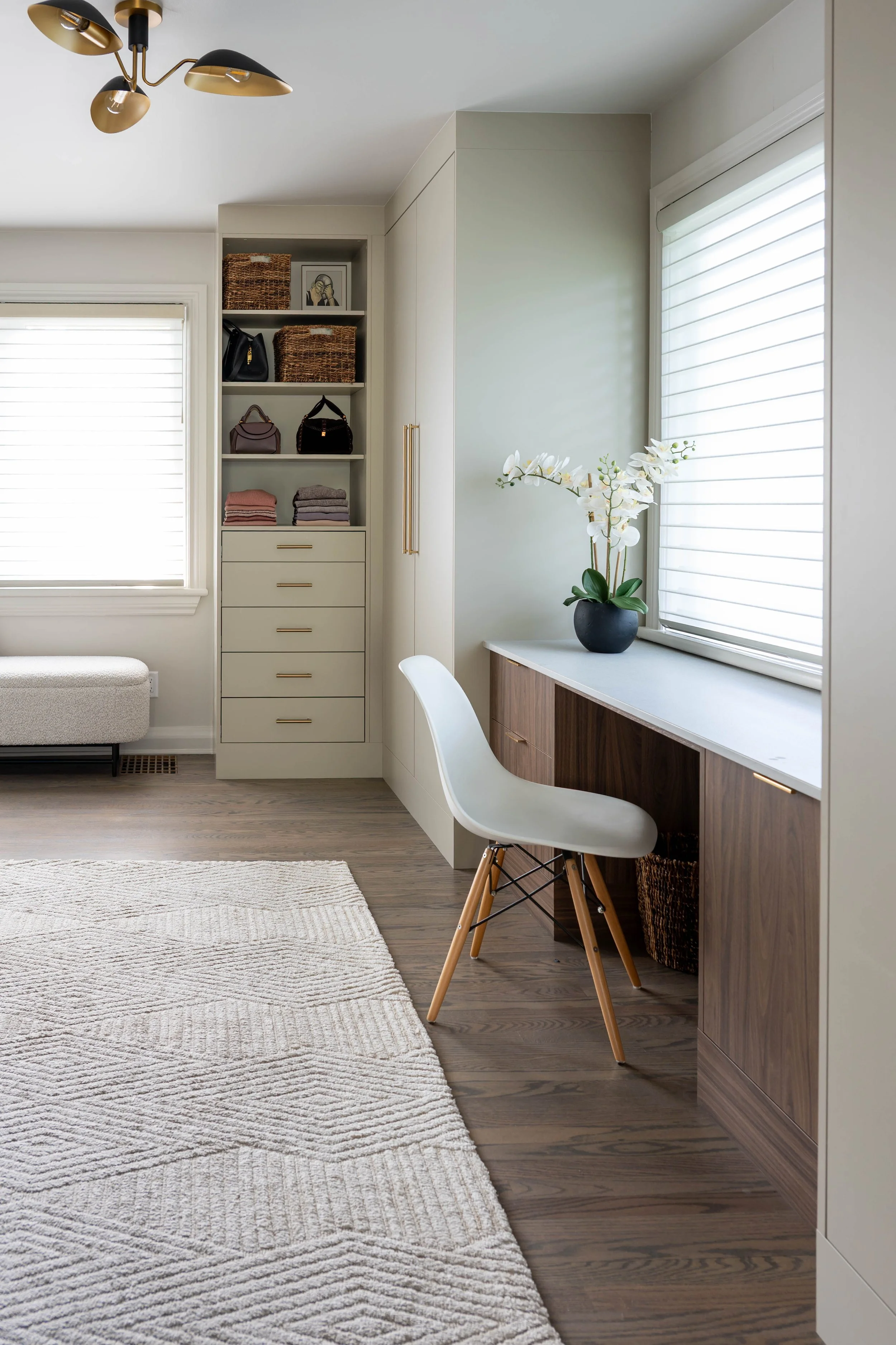 An interior room featuring a built-in desk with a white chair, a black vase with white orchids, a window with white blinds, cabinetry with gold handles, and a white and beige rug on wooden flooring.