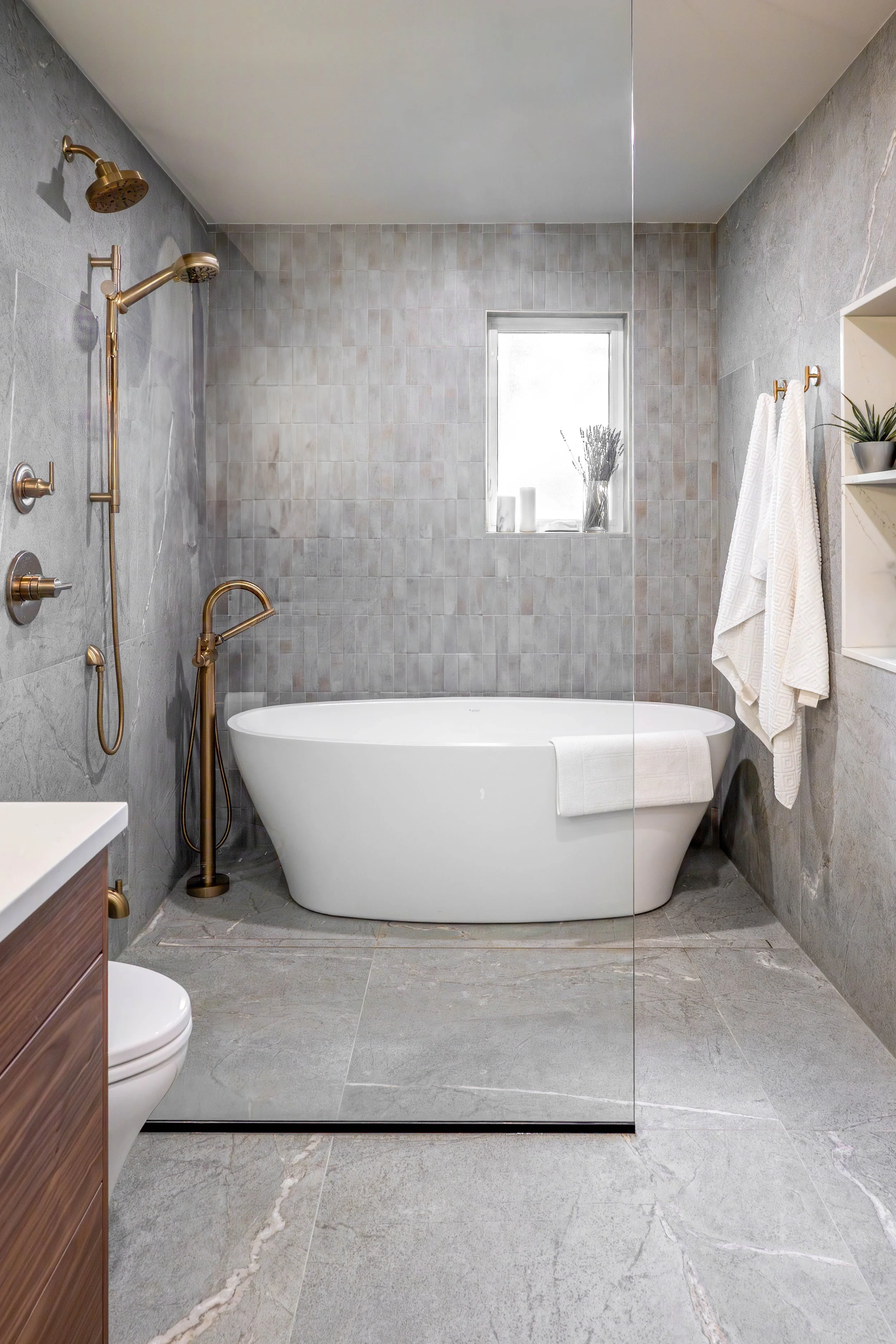 Modern bathroom with a freestanding bathtub, gray tiled walls and floor, a small window, and brass fixtures, including a shower head and faucet.