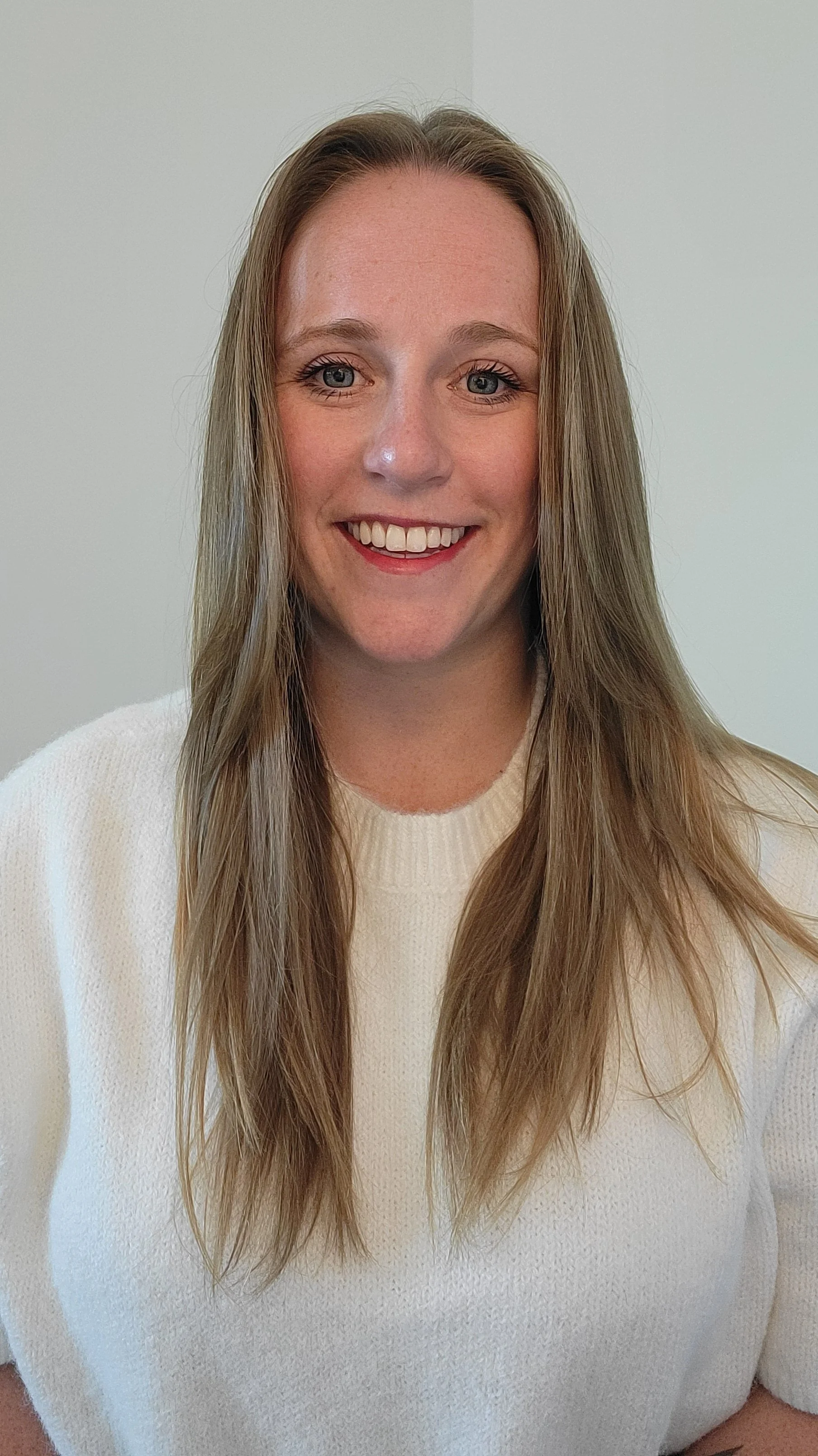 A woman with light skin, brown hair that is straight and long,, wearing a white sweater, gold hoop earrings, and bright lipstick, smiling at the camera.