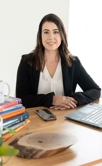A woman with dark brown hair wearing a black blazer and white top sitting at a wooden desk with a silver laptop, a smartphone, a stack of books, a mug, and a wooden sign that reads 'MINDSHOP'.