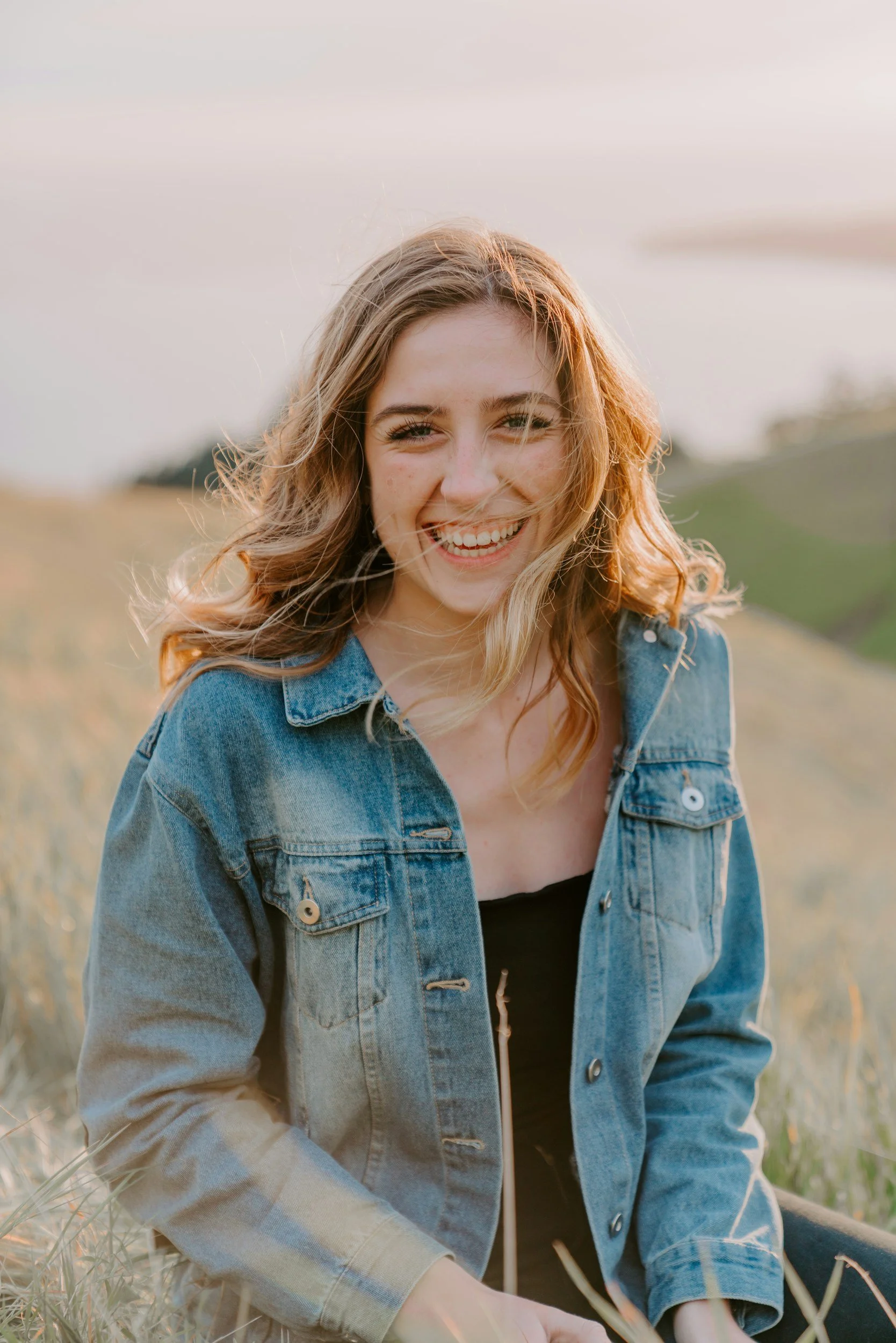 A young woman with wavy hair sitting outdoors, smiling at the camera, wearing a denim jacket.