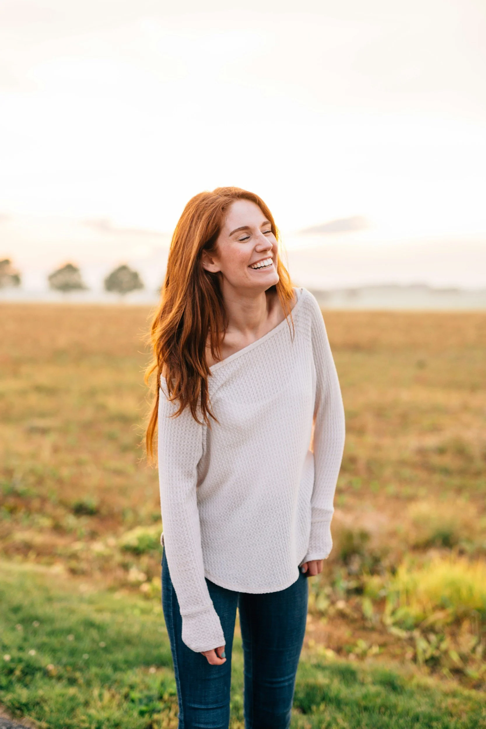 A woman with red hair smiling outdoors in a field during sunset.