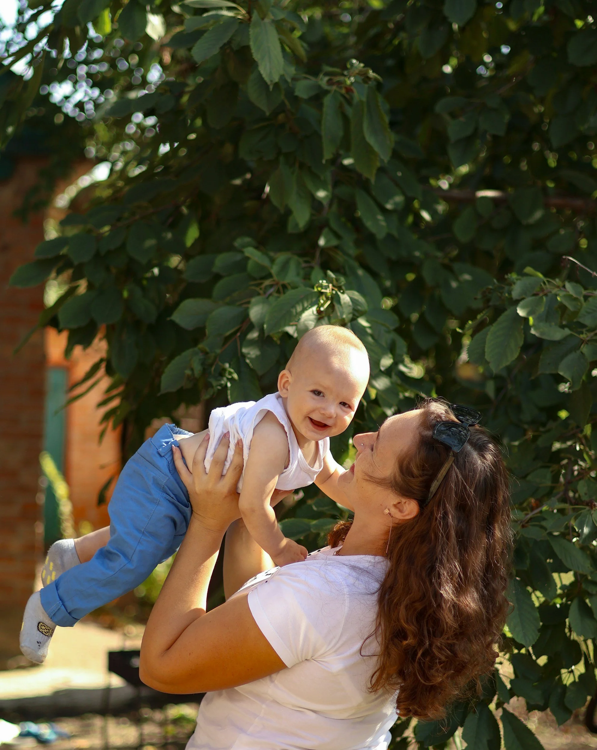 A woman with curly hair lifting a baby boy with a big smile in front of green leafy trees outdoors.
