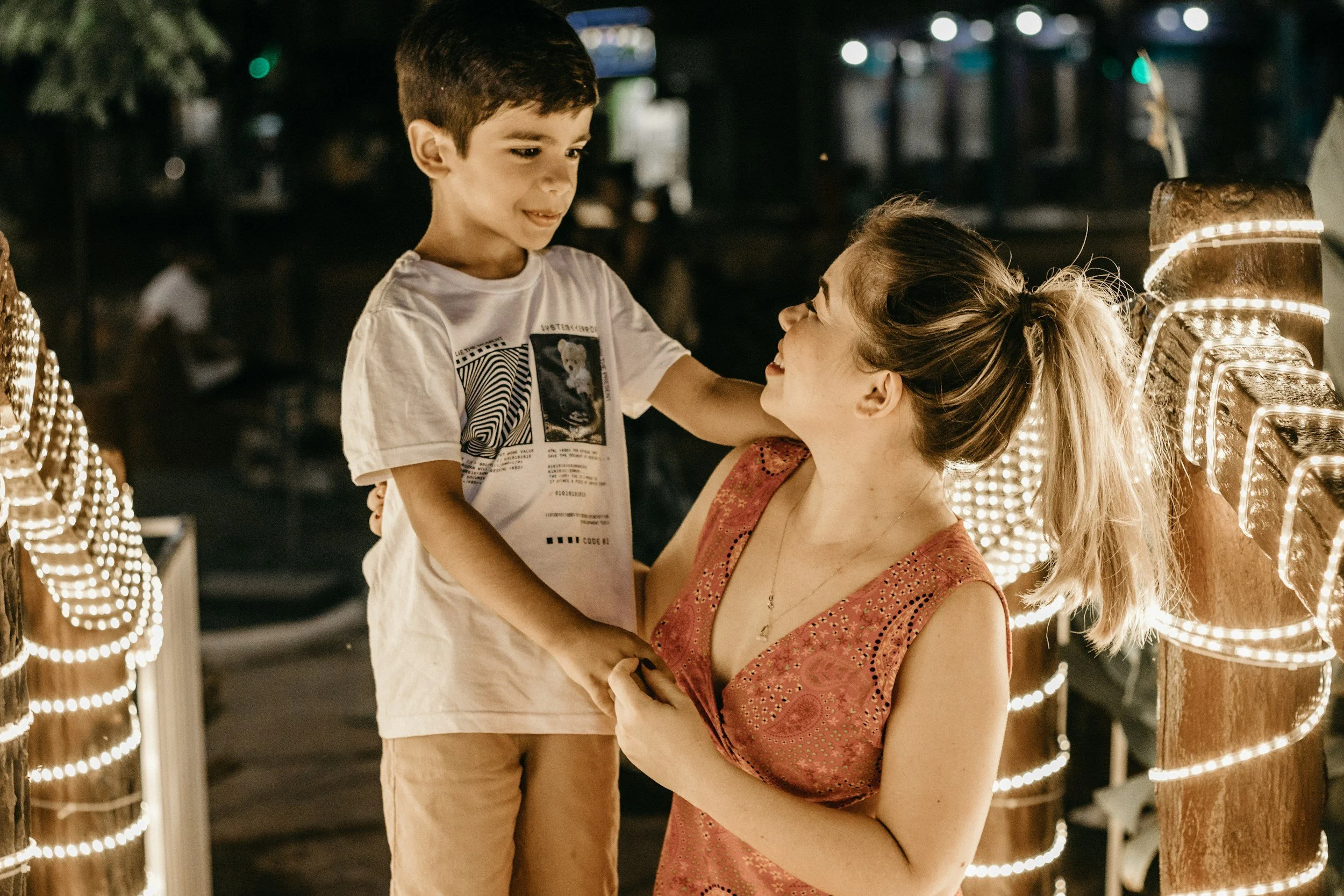 A woman lifting a young boy with short dark hair and wearing a white T-shirt, at night with illuminated decorative lights around them, smiling and making eye contact.