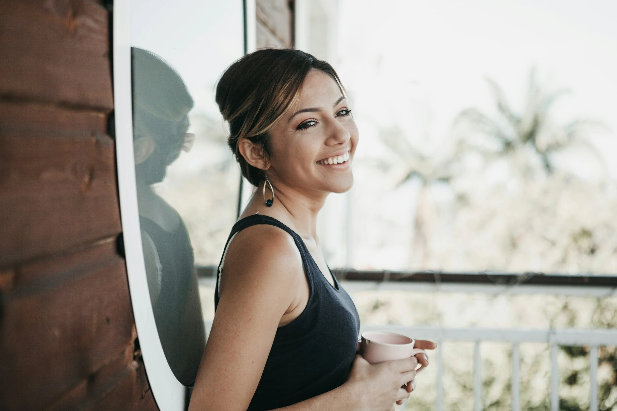 A smiling woman with short hair, earrings, and a black sleeveless top, holding a pink mug, standing on a balcony with trees in the background.
