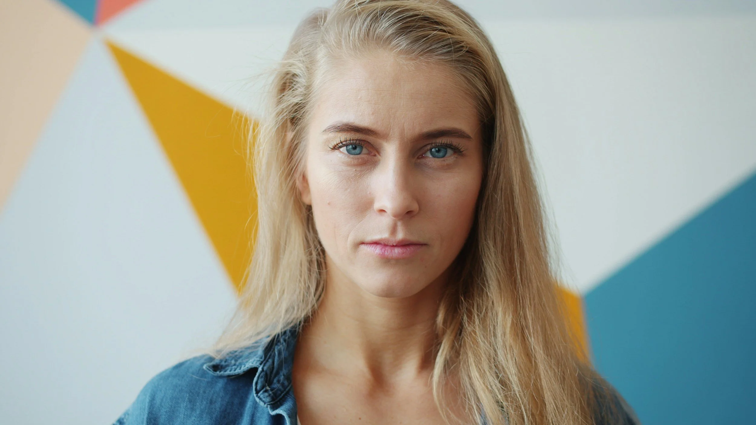 A young woman with blonde hair and blue eyes standing in front of a wall painted with a geometric pattern of blue, yellow, and white shapes.