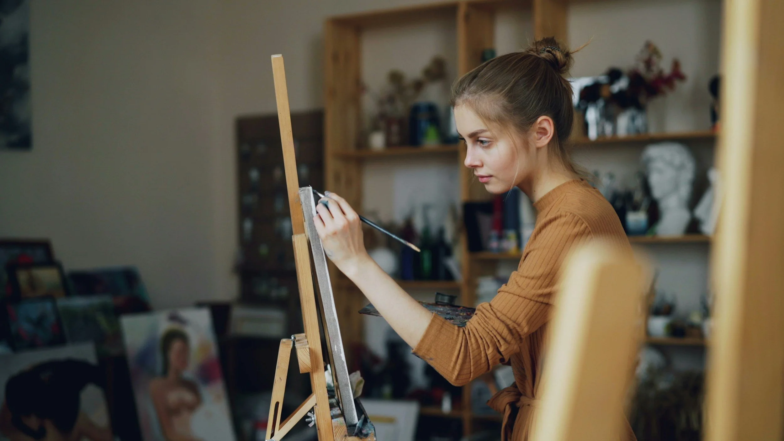 Young woman painting on a canvas in an art studio with shelves of art supplies and artwork in the background.