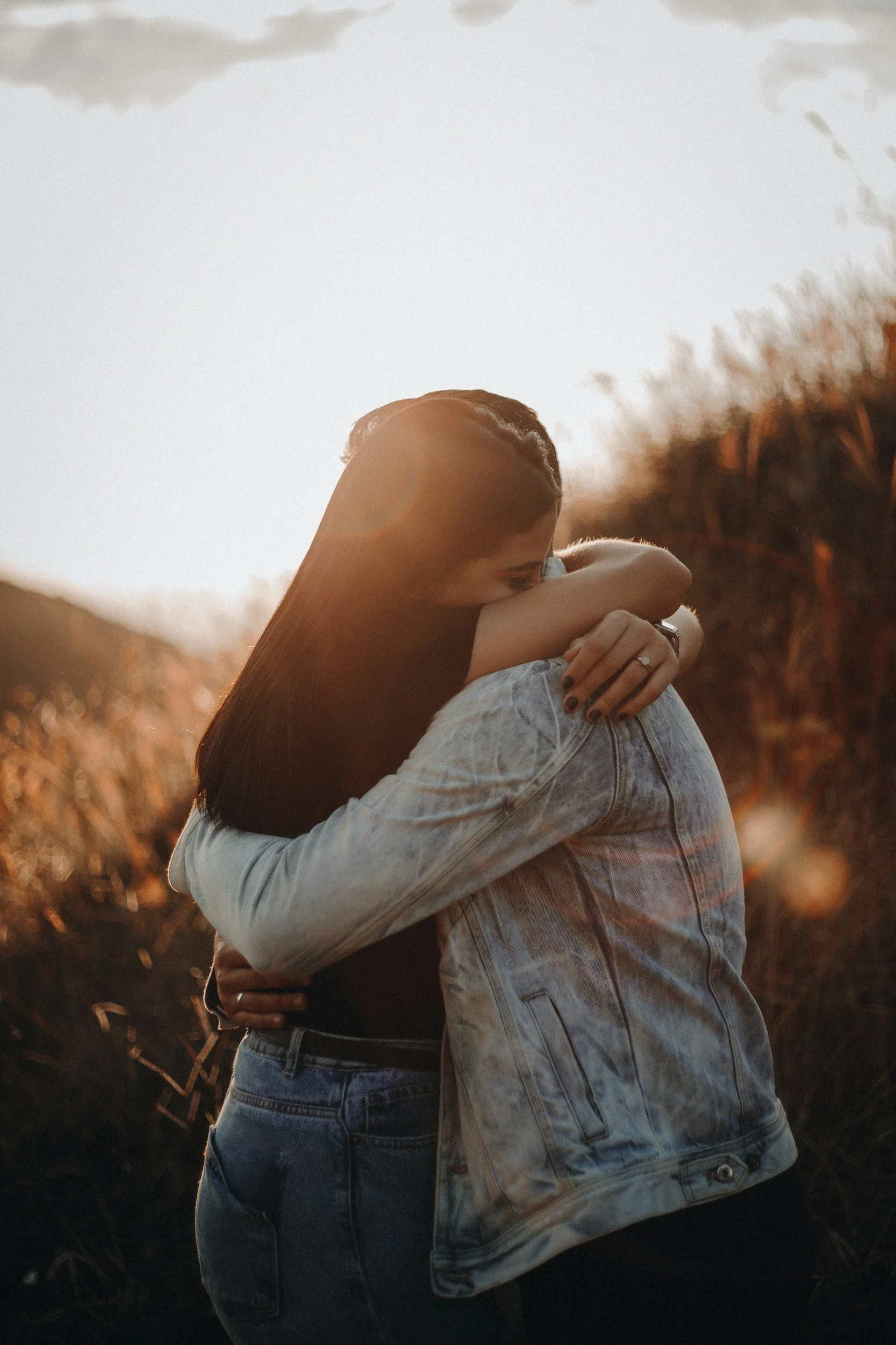 Two women embracing outdoors during sunset, one with braided hair and the other with long straight hair, in a field with tall dry grass.