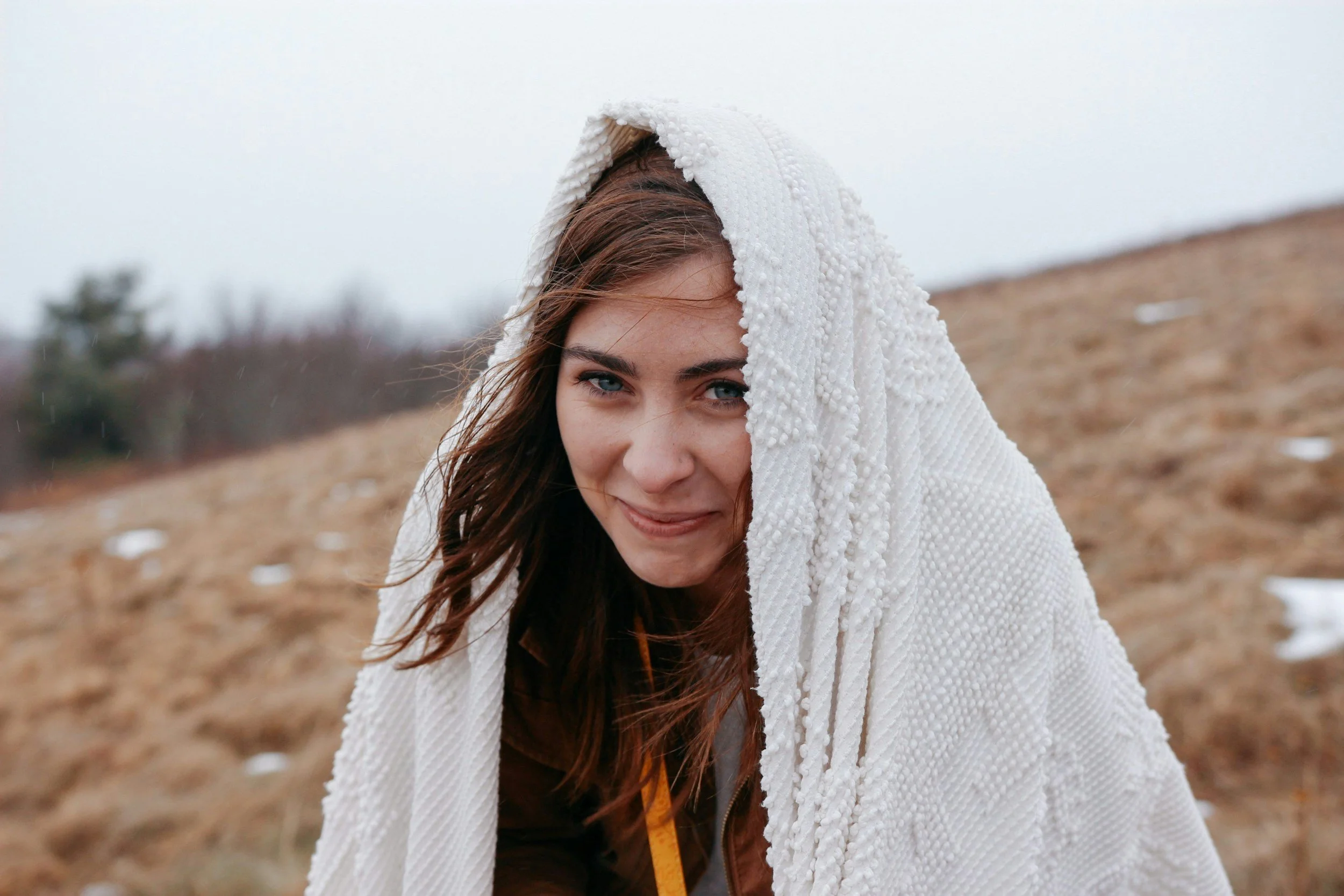 Young woman with brown hair and blue eyes smiling outdoors in a field, wrapped in a white textured blanket or shawl, with cloudy sky in the background.
