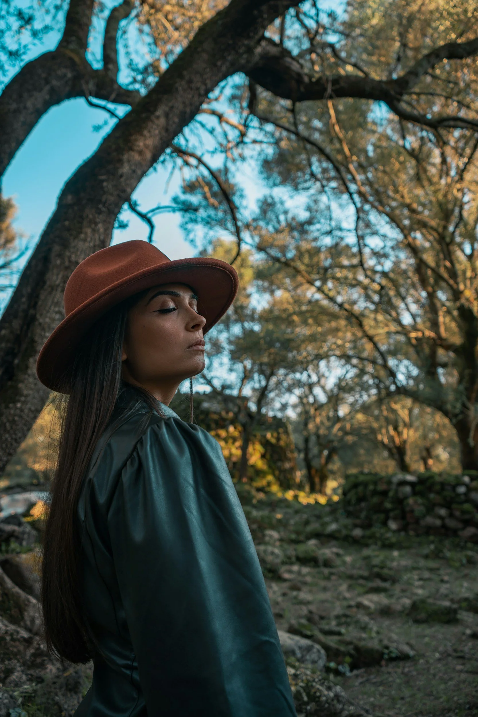 A young woman with long dark hair wearing a brown wide-brimmed hat, a black jacket, and closed eyes, stands outdoors among trees with sunlight filtering through the branches.