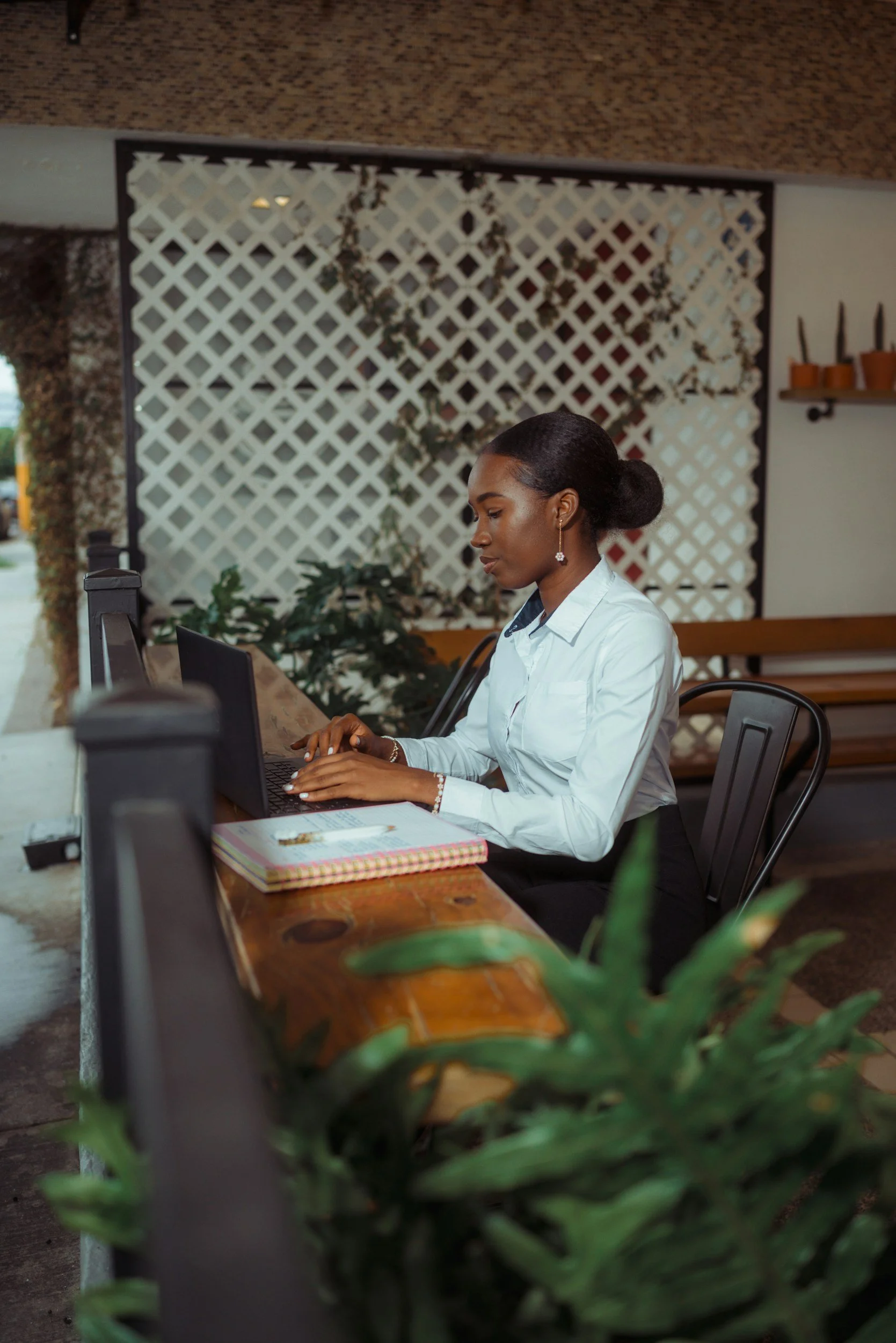 A woman sitting at a wooden table working on a laptop, with notebooks nearby in a cozy cafe or workspace with plants and decorative wall lattice.