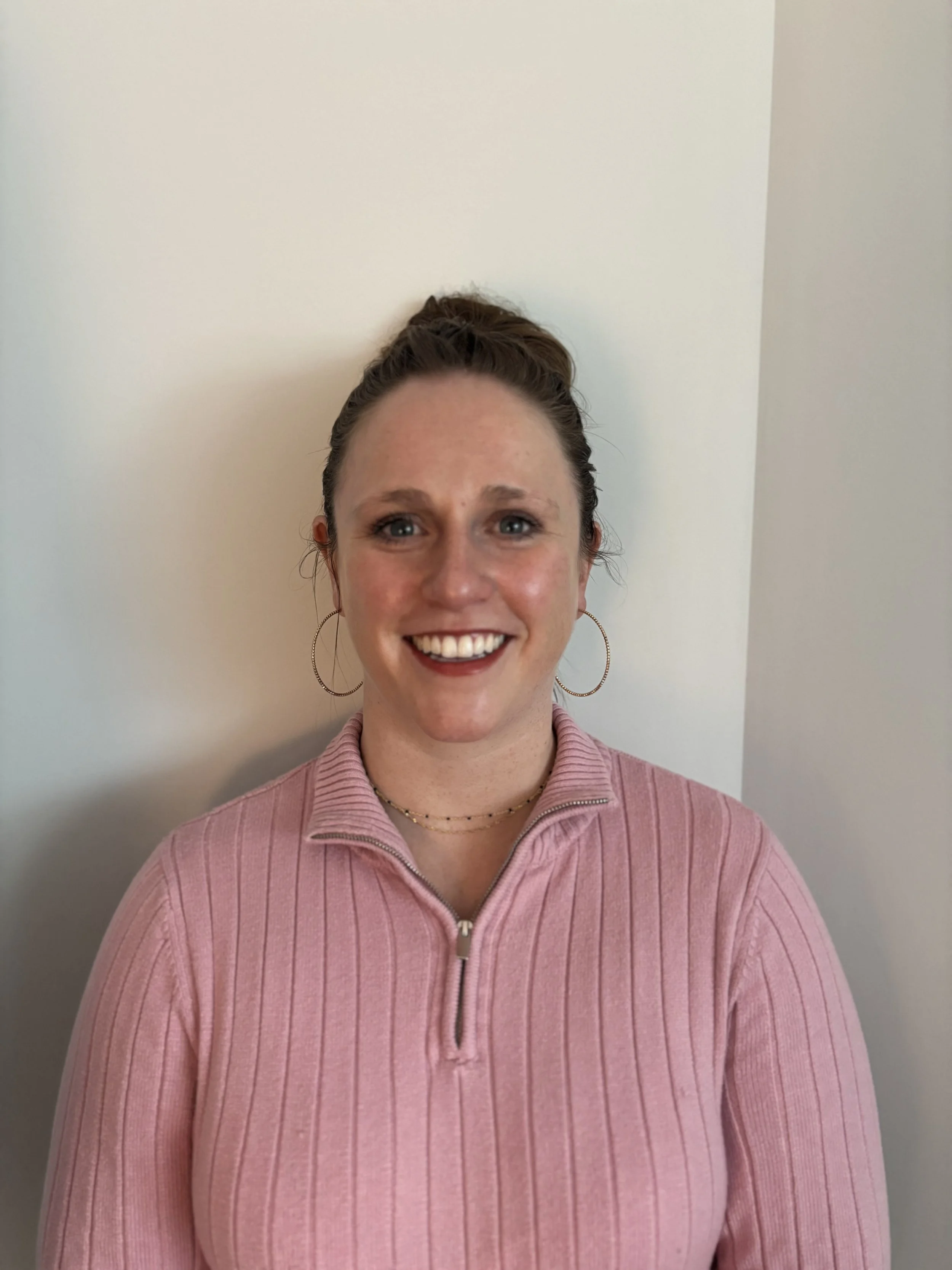 A woman with light skin, brown hair in a high bun, wearing a pink zip-up sweater, gold hoop earrings, and bright lipstick, smiling at the camera.