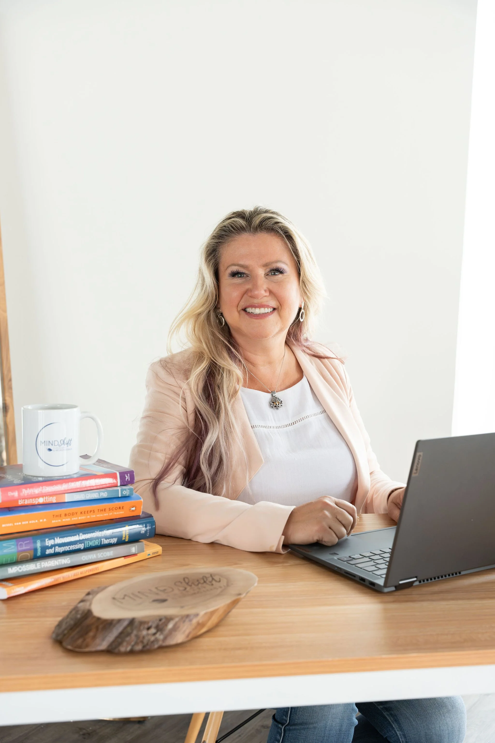A women sitting at a wooden desk, with a laptop, with text books and a mug, she is working as a counsellor and psychotherapist