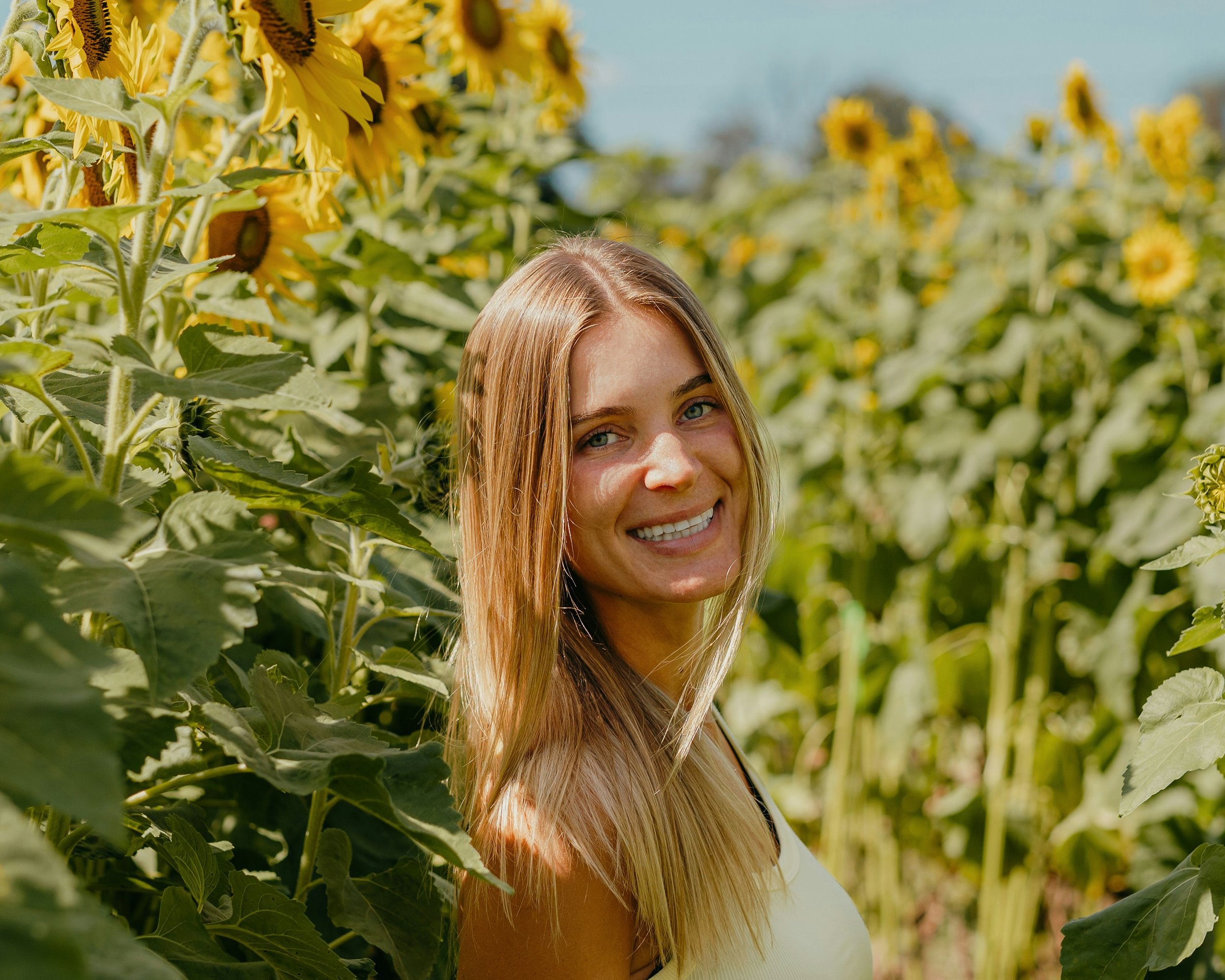 A young woman with long blonde hair smiling in a sunflower field on a sunny day.