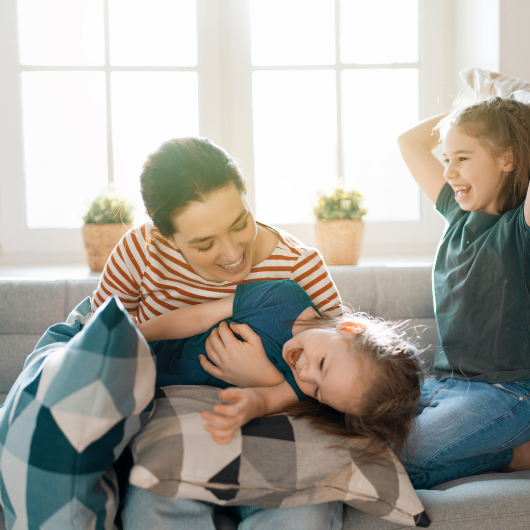 A woman and two children playfully wrestling and laughing on a couch in a bright room with large window and potted plants.