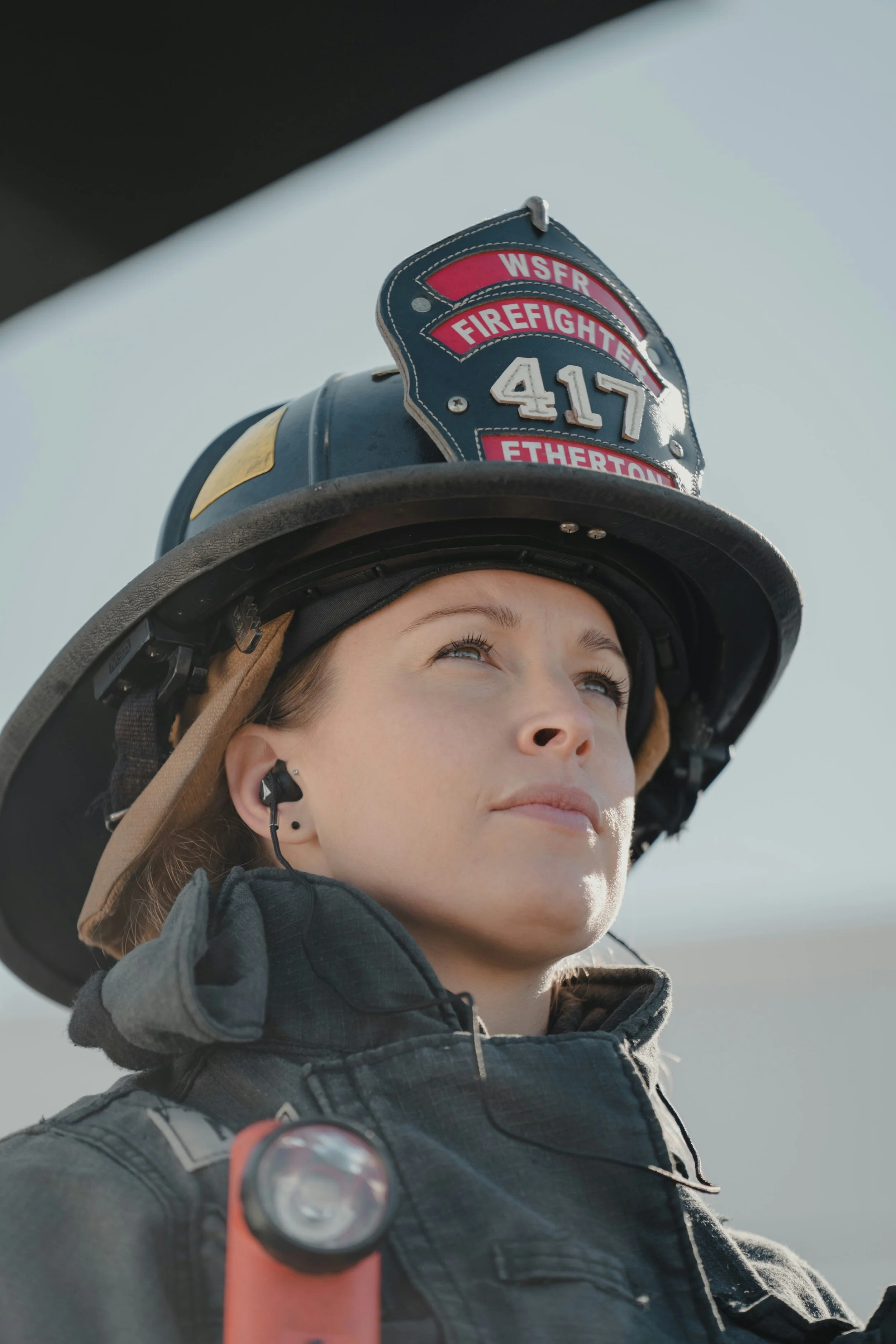 A female firefighter wearing a helmet with a badge labeled 'WSFR Firefighting 417 Etherton' and an earpiece, looking up with sunlight on her face.