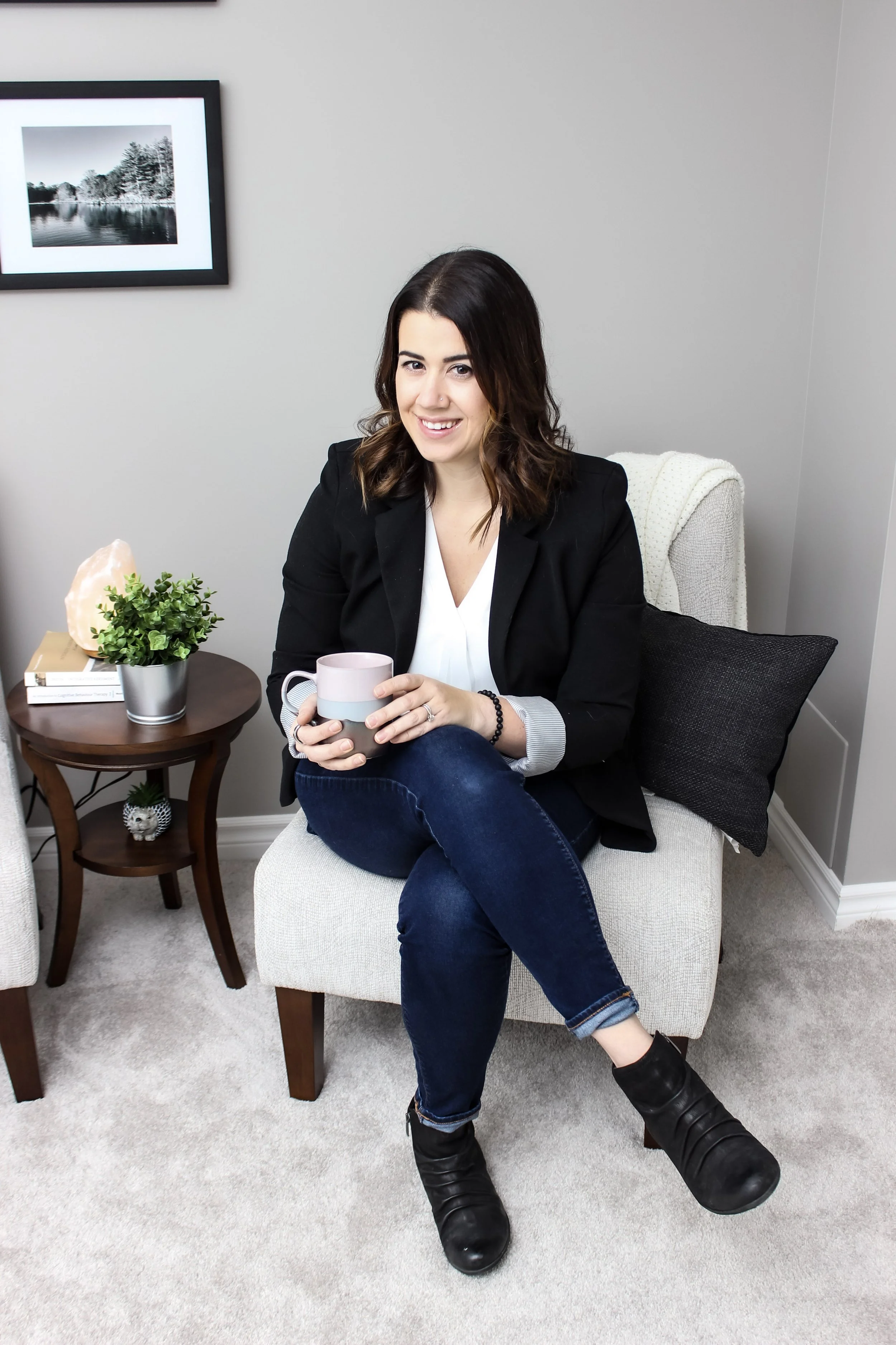 A woman with shoulder-length brown hair, dressed in a black blazer, white shirt, and dark jeans, sitting on a beige armchair in a living room, holding a pink mug, and smiling at the camera.