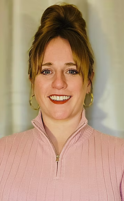 A woman smiling, with her hair styled in an updo, wearing a pink zip-up top and gold earrings, standing in front of a neutral background.