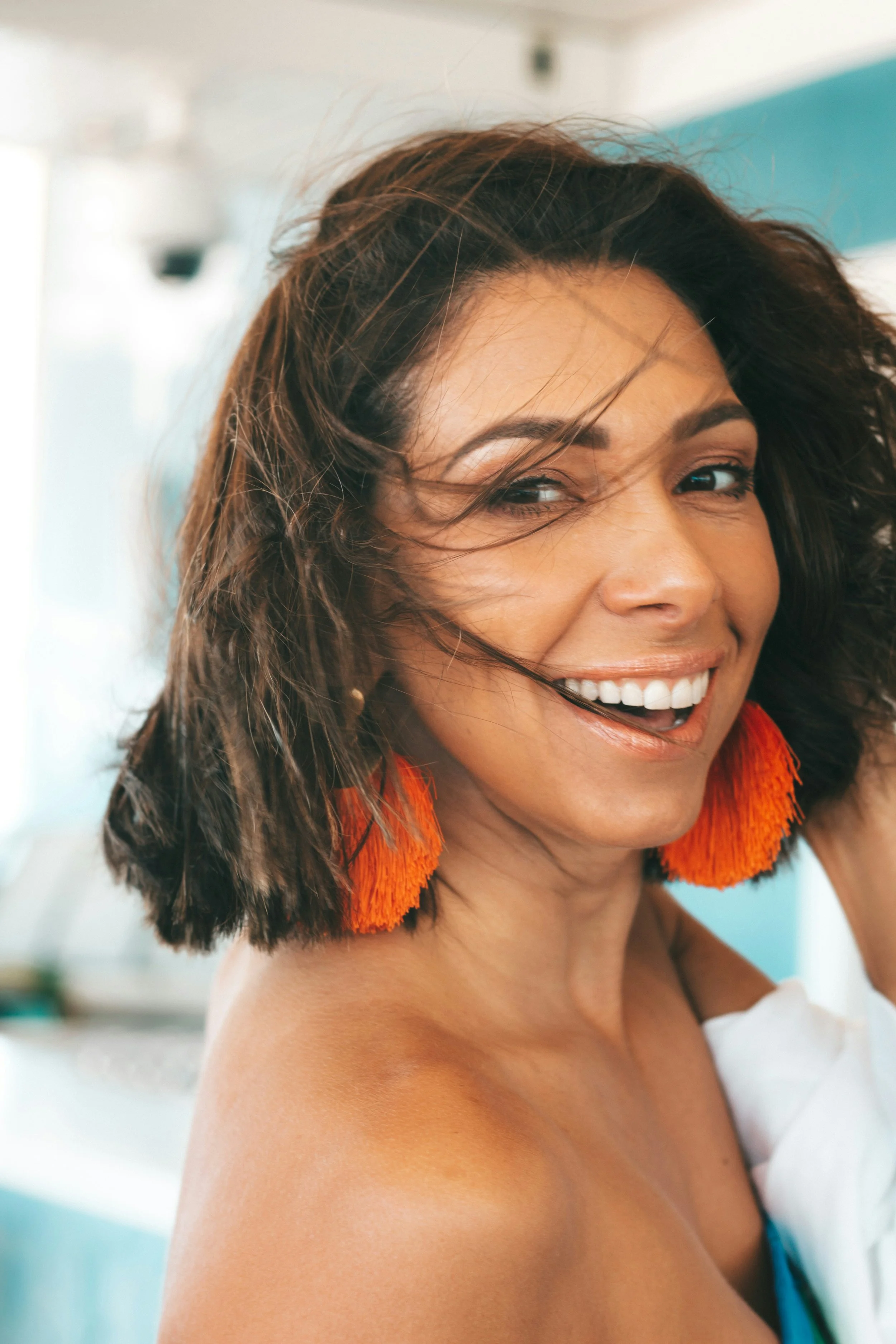 Close-up of a smiling woman with shoulder-length dark hair, wearing large orange tassel earrings, in a bright setting, possibly outdoors or near a window.