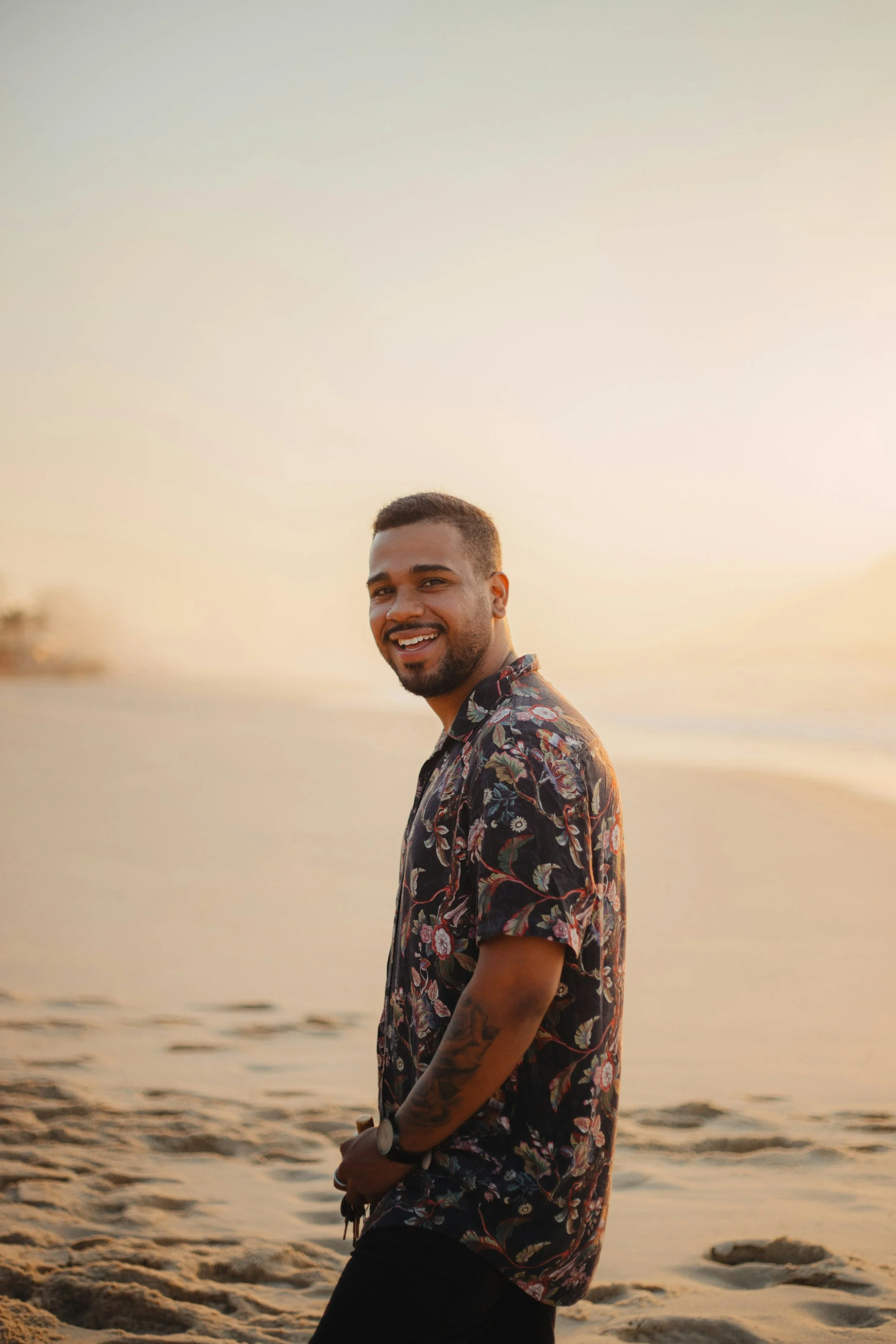 Smiling man in a floral shirt standing on the beach at sunset.