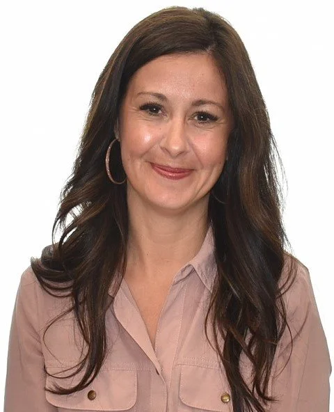 A woman with long dark brown hair wearing a light pink button-up shirt and large hoop earrings smiling at the camera.