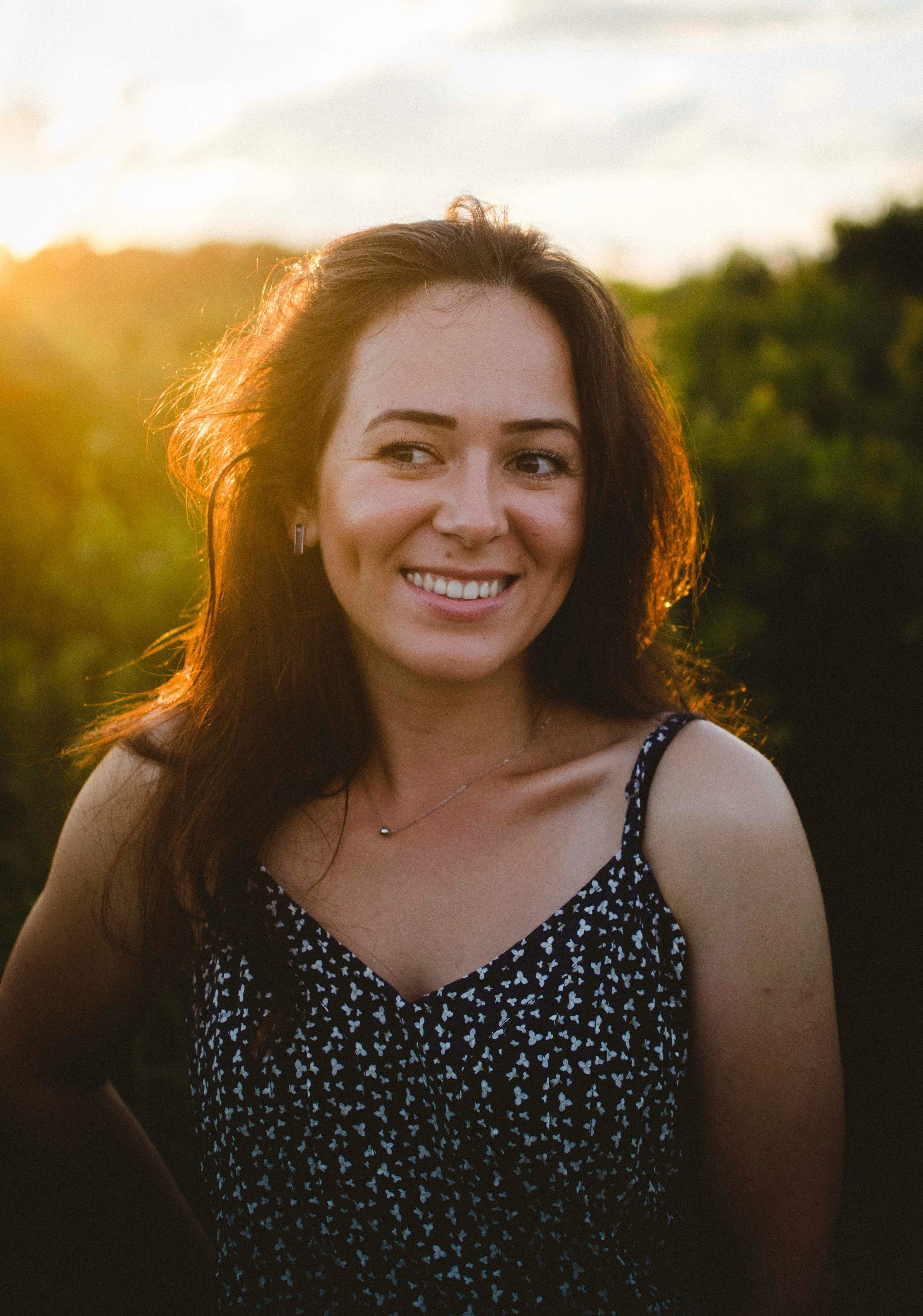 A woman with dark brown hair smiling outdoors during sunset with greenery in the background, wearing a black dress with white floral print and a silver necklace.
