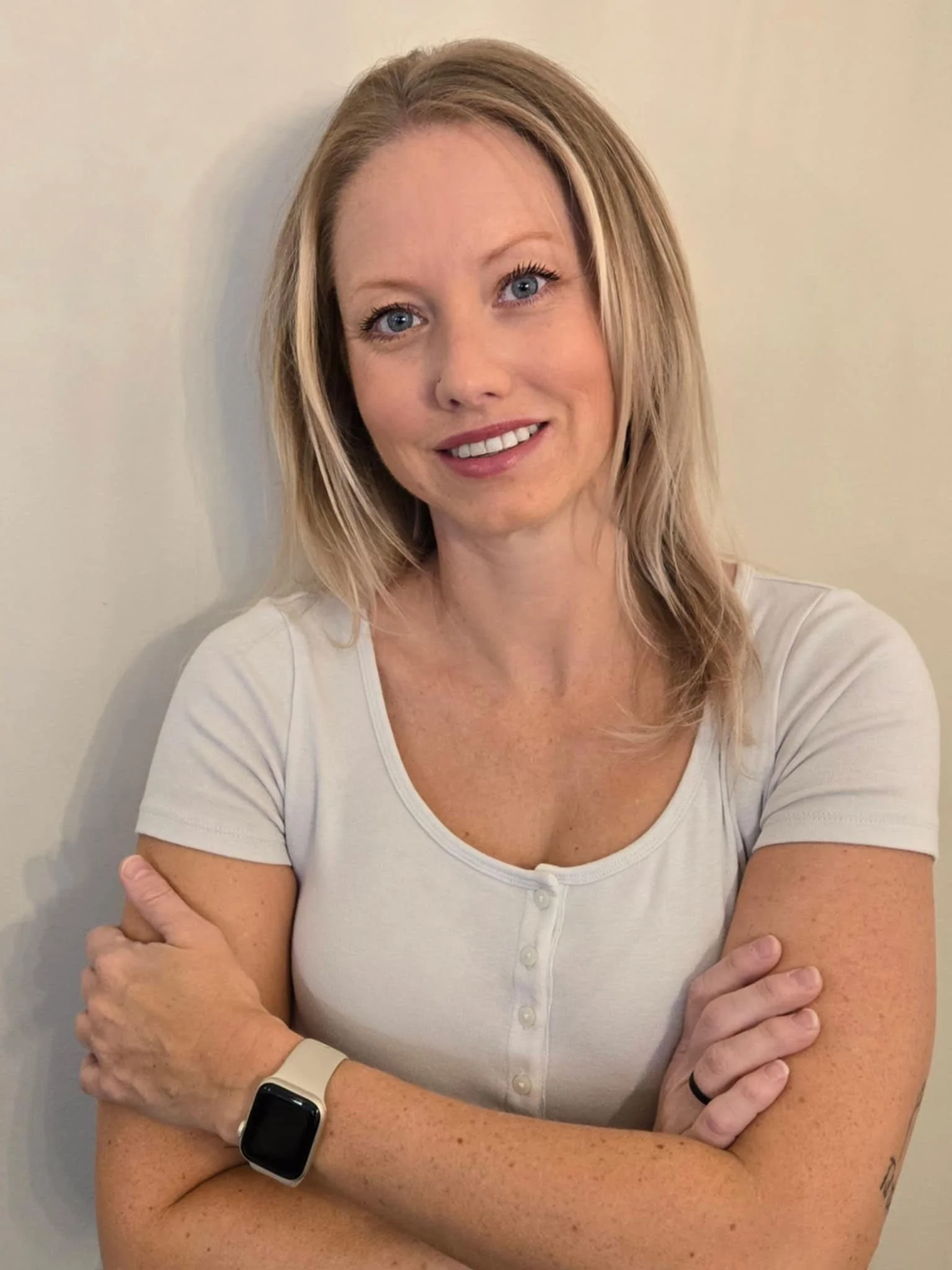 A woman with shoulder-length blonde hair and blue eyes smiling, wearing a light-colored, short-sleeved, button-down shirt and a smartwatch on her left wrist, standing against a plain wall.