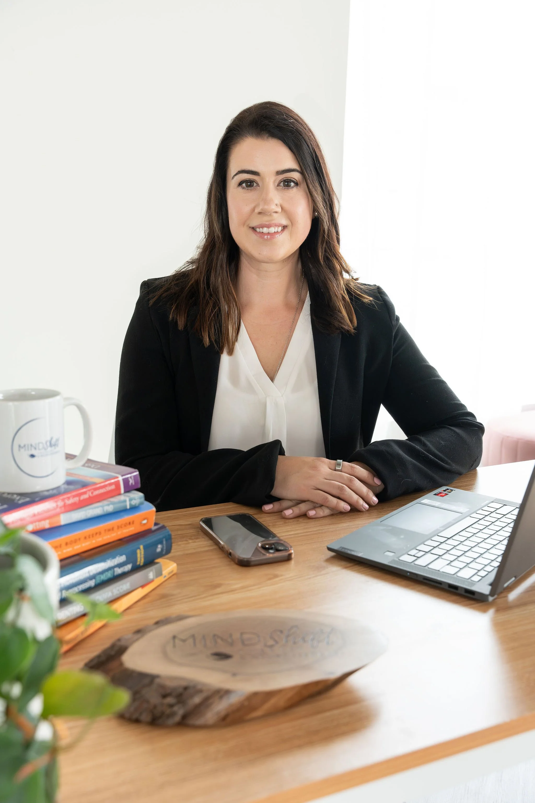 A woman with dark brown hair sitting at a wooden desk with a laptop, a smartphone, a mug, and a stack of books. There is a blurred plant in the foreground and a white background.
