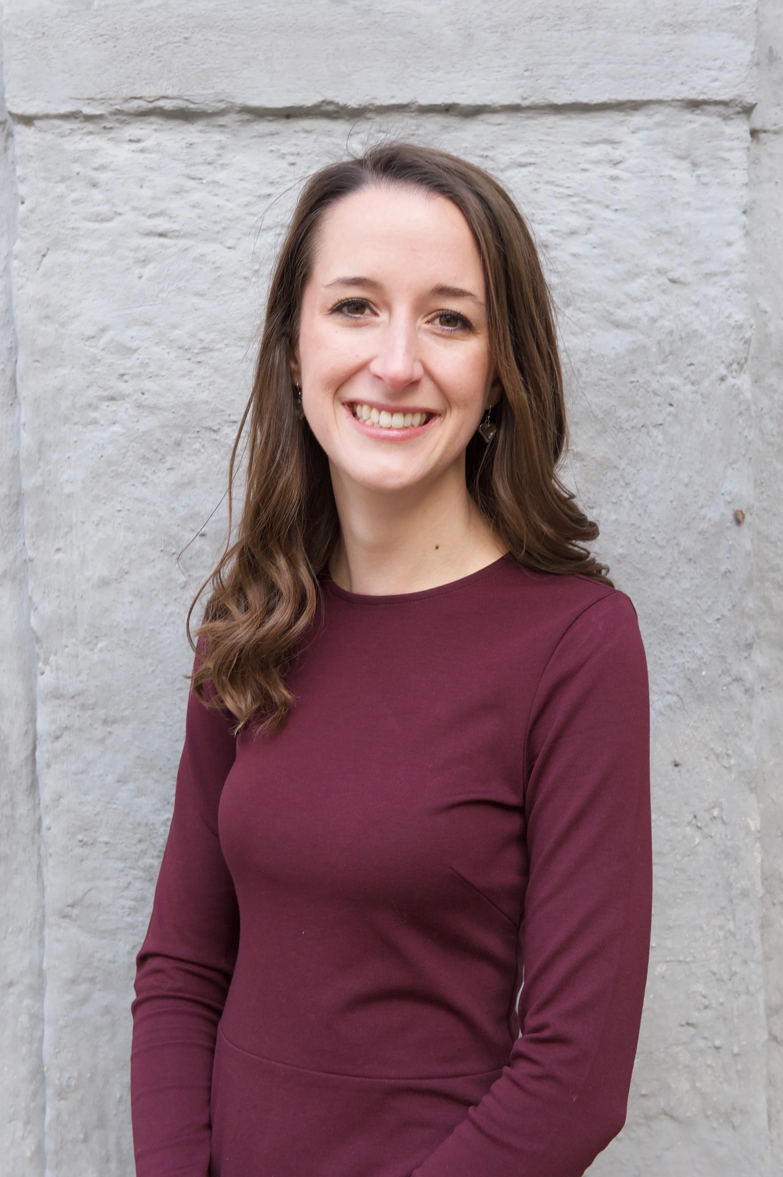A smiling woman with long brown hair wearing a maroon long-sleeve shirt, standing outdoors against a light gray stone wall.