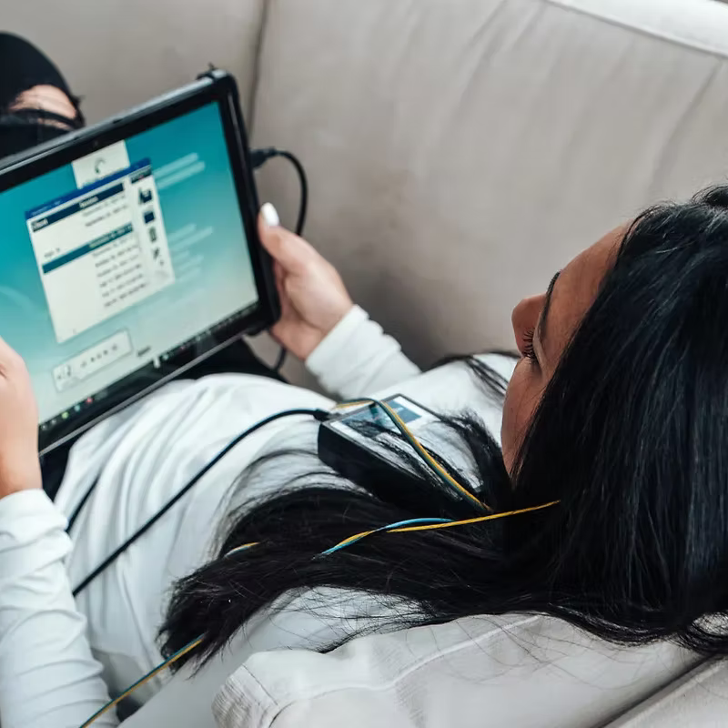 A woman with long black hair lying on a beige couch, watching a tablet screen, with earbuds connected, and her head resting on a white shirt.