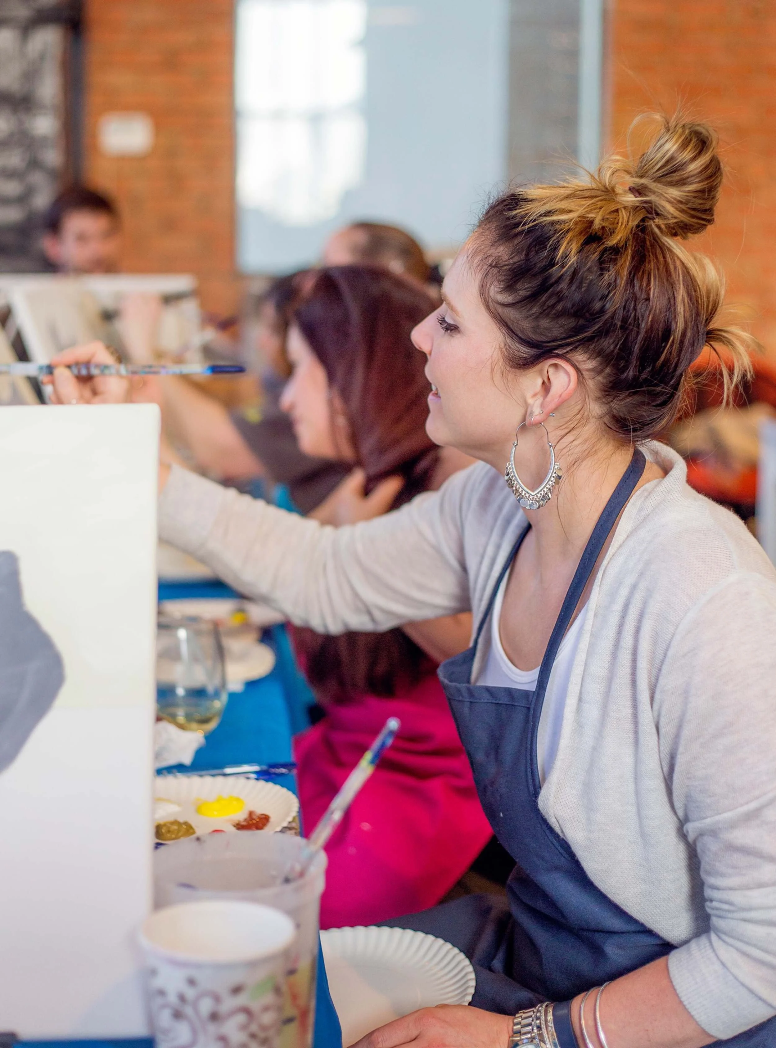 Women participating in a painting class, focusing on their canvases, with art supplies and drinks on the table.