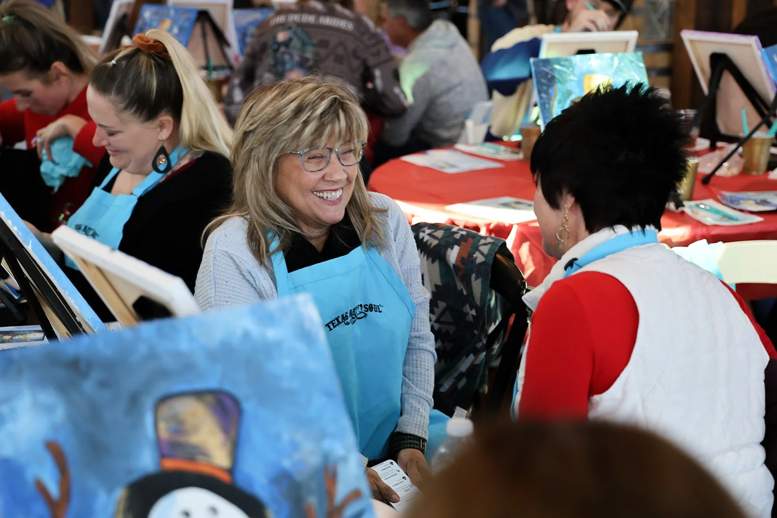 Two women are talking and smiling at a painting event, with other participants in the background. They are wearing aprons and appear engaged in conversation.